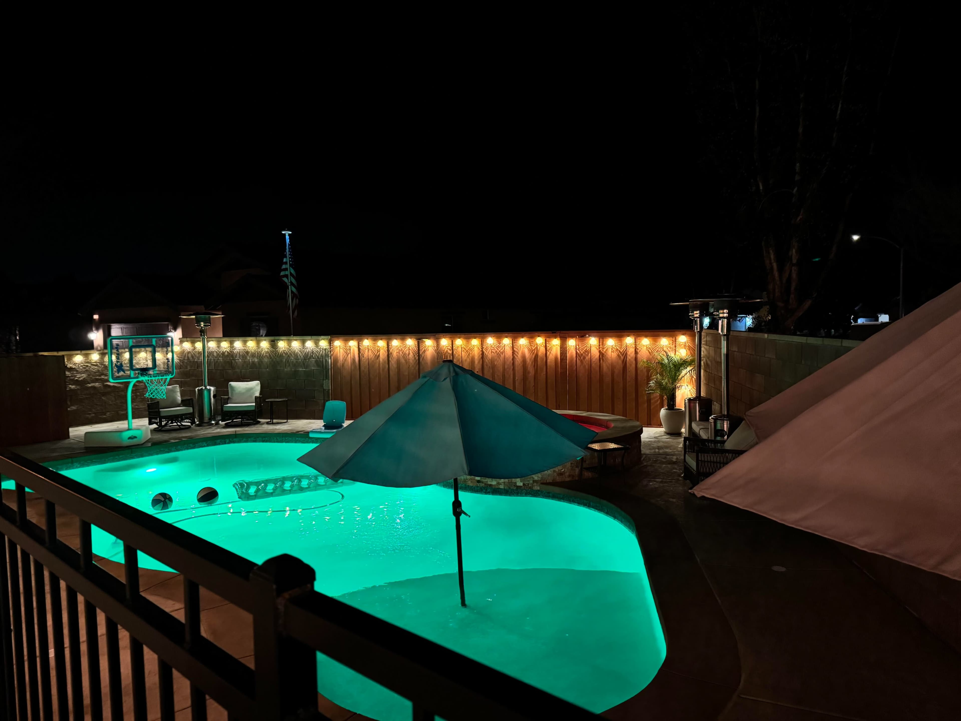 The image shows a lit swimming pool at night, surrounded by lounge chairs and a patio area with an umbrella and a tent.