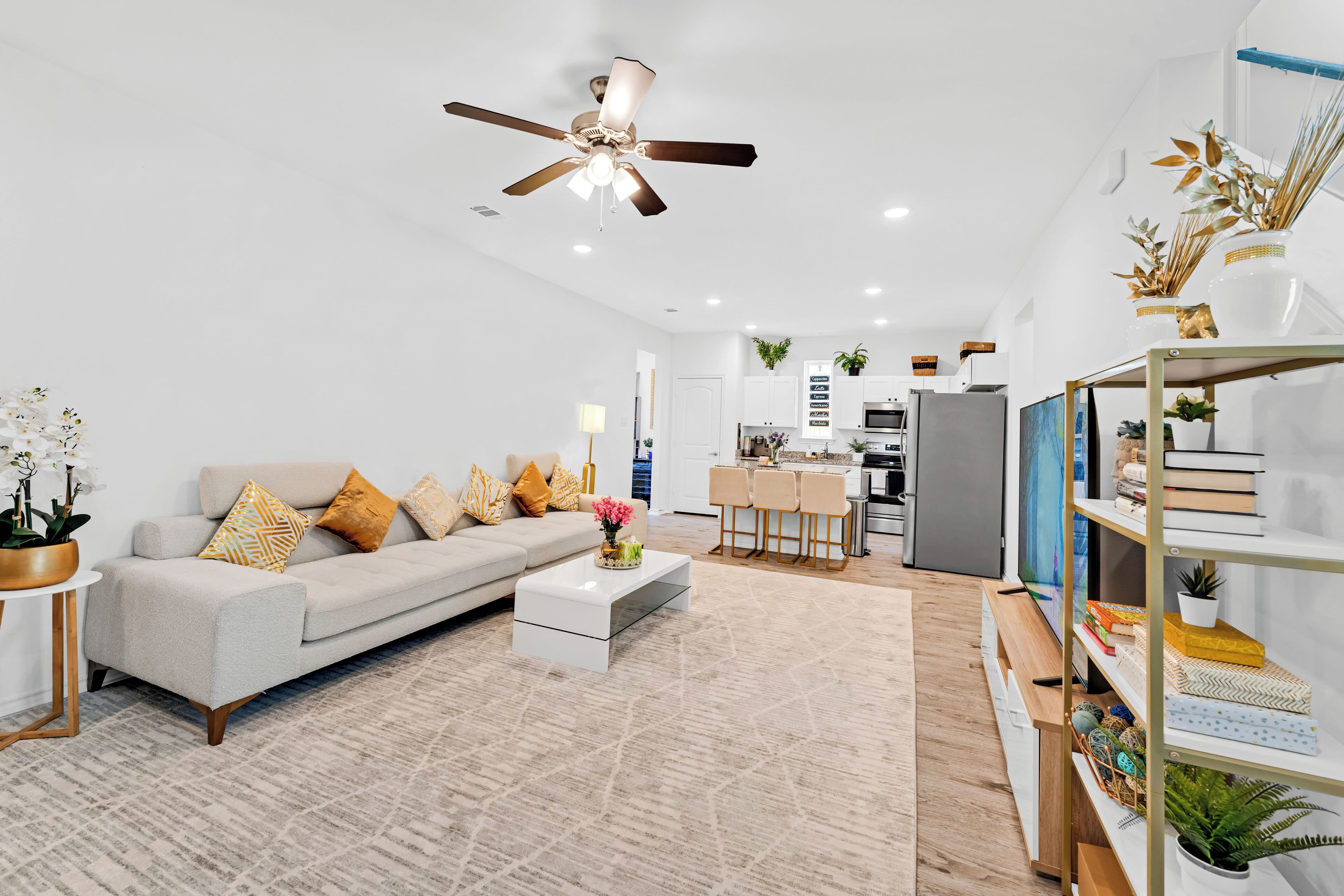 A modern living room with a light-colored sofa, a coffee table, and a dining area visible in the background, featuring a kitchen and decorative shelving.
