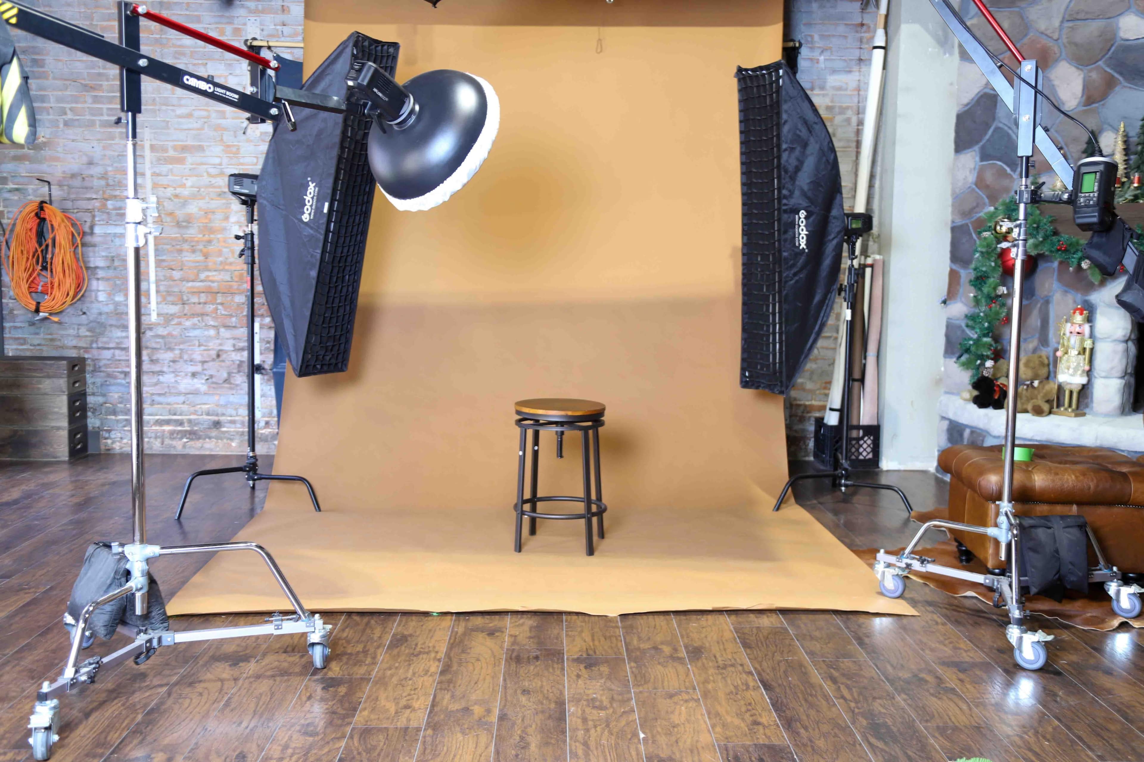A photography studio setup featuring a brown backdrop, a wooden stool, and two softbox lights positioned on either side.