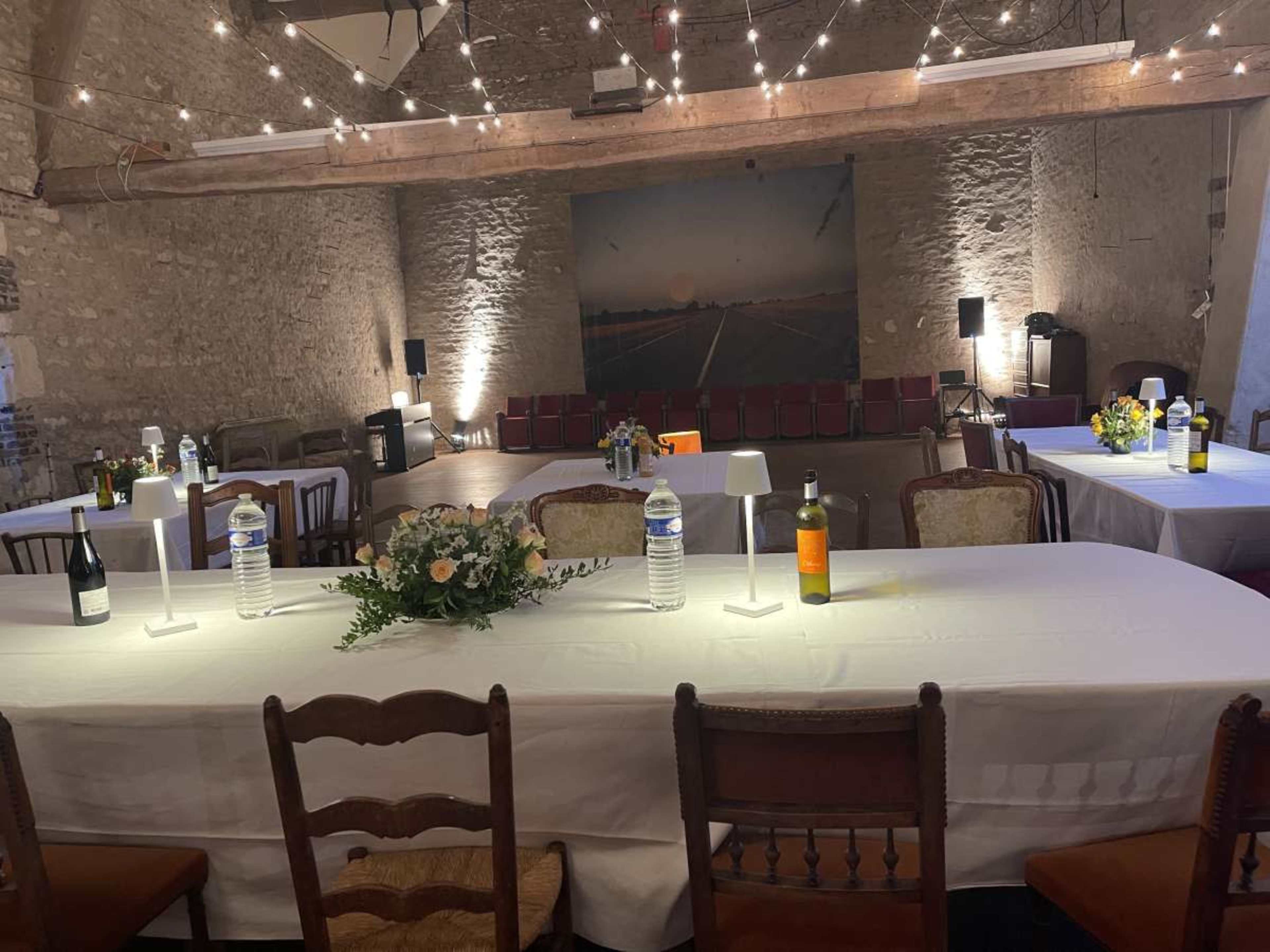 A spacious event room features several tables set with bottles and floral centerpieces, surrounded by chairs and illuminated by string lights.