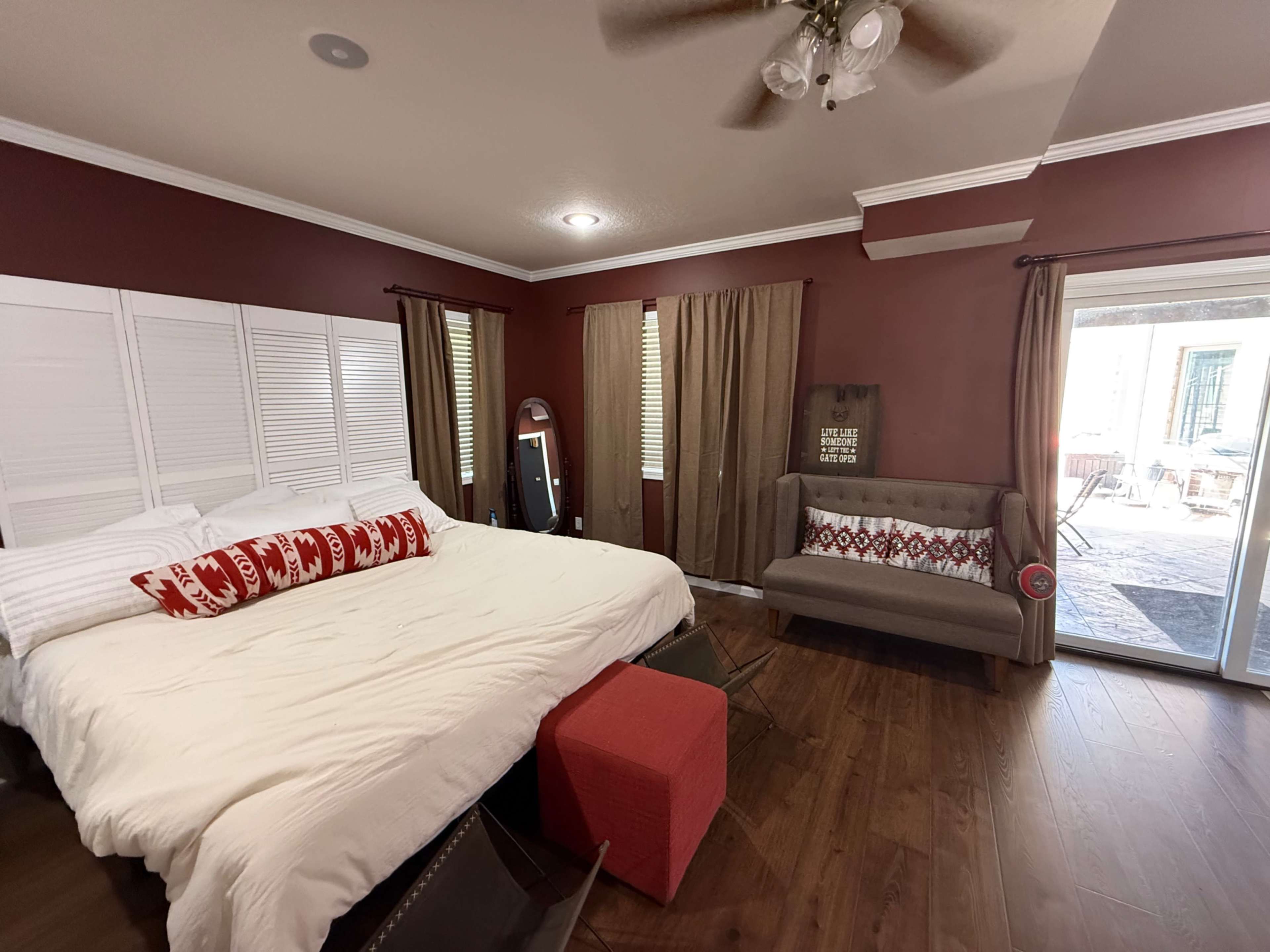 The image shows a bedroom with a large bed, a couch, and sliding glass doors leading to an outdoor area, all set against a deep red wall.
