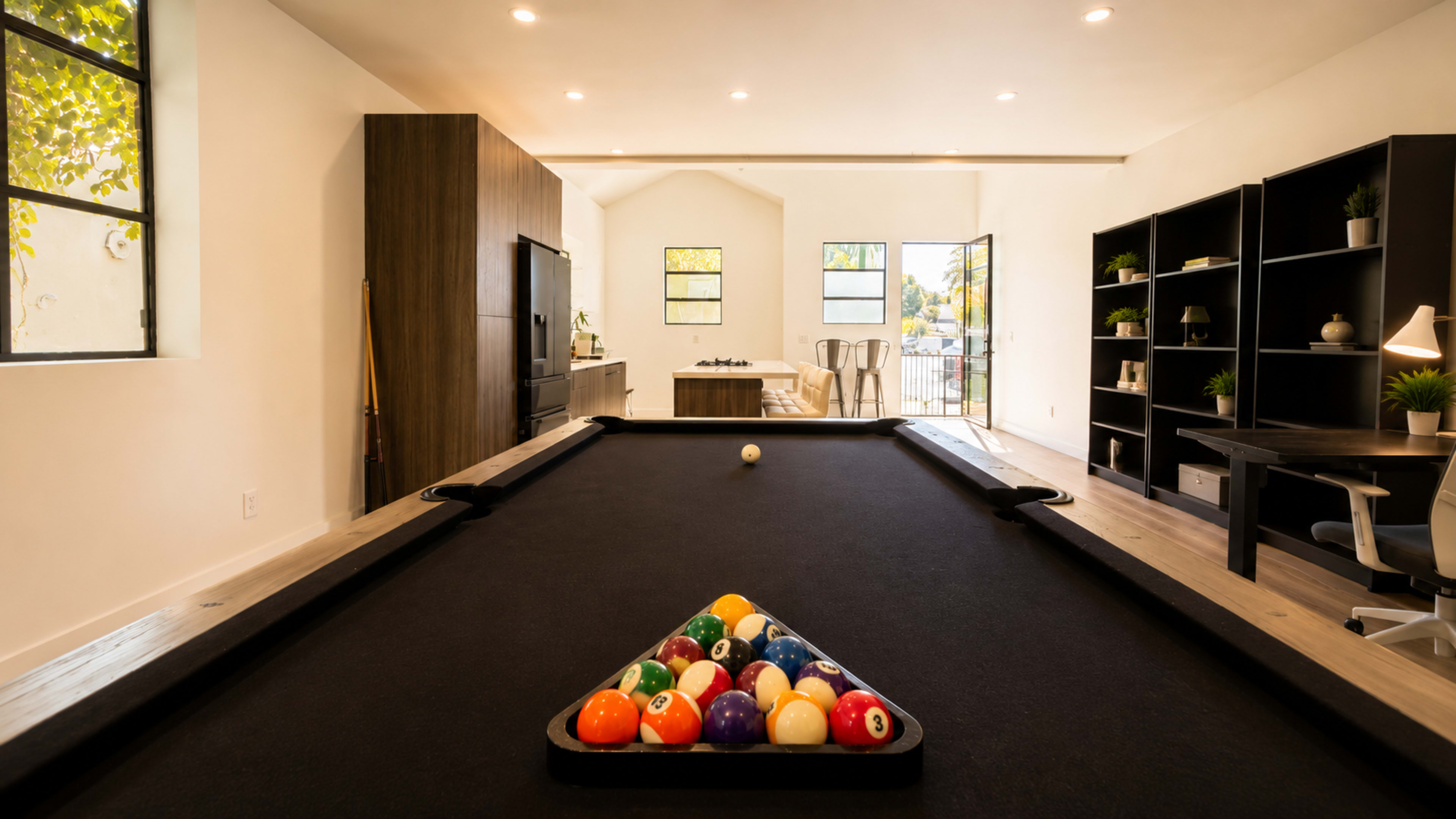 A modern game room features a black pool table with a triangle of colorful billiard balls and a cozy seating area with shelves and a large window.