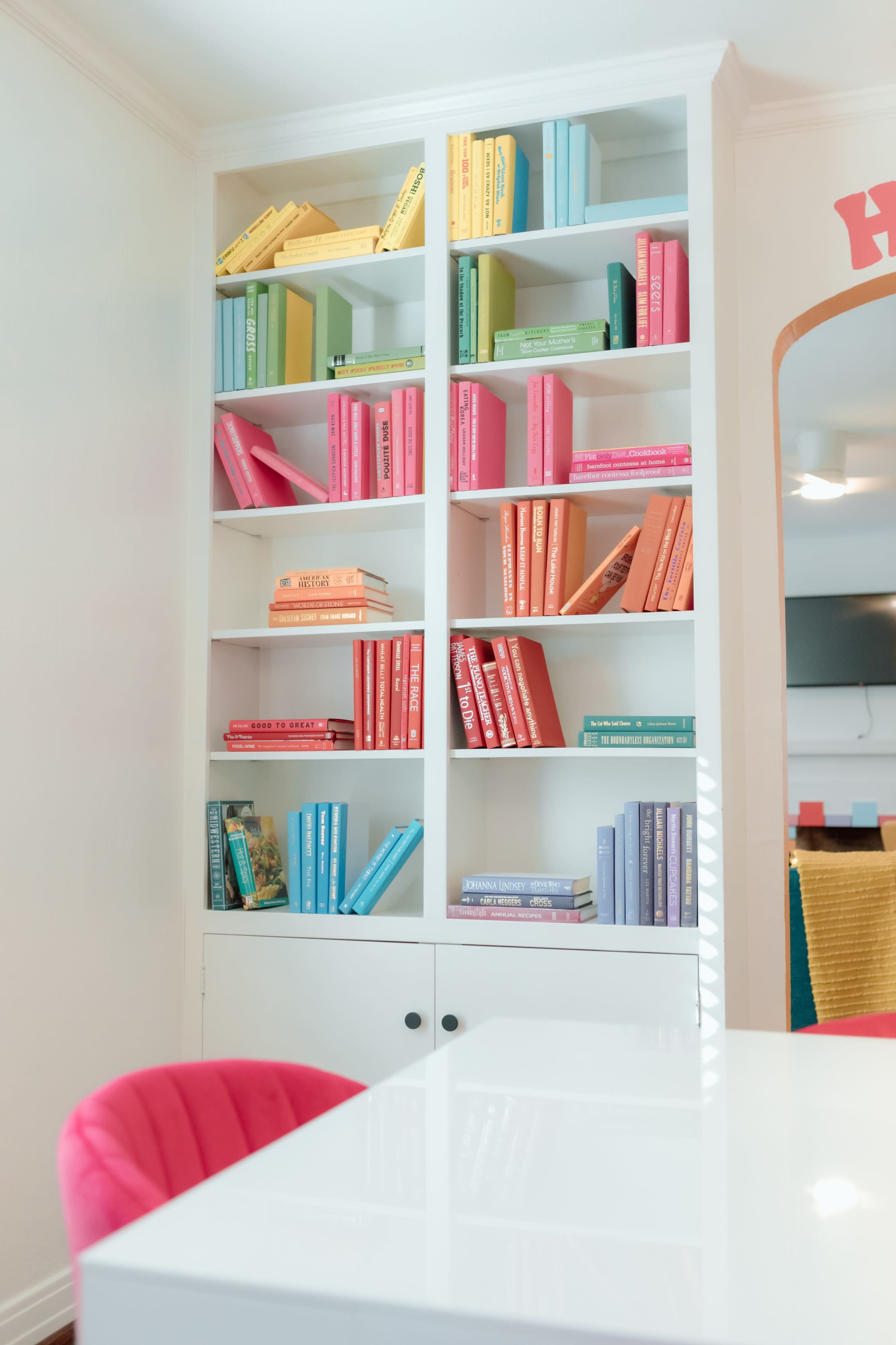 A white bookshelf filled with colorful books is set against a light wall in a bright room, accompanied by a pink chair and part of a table in the foreground.