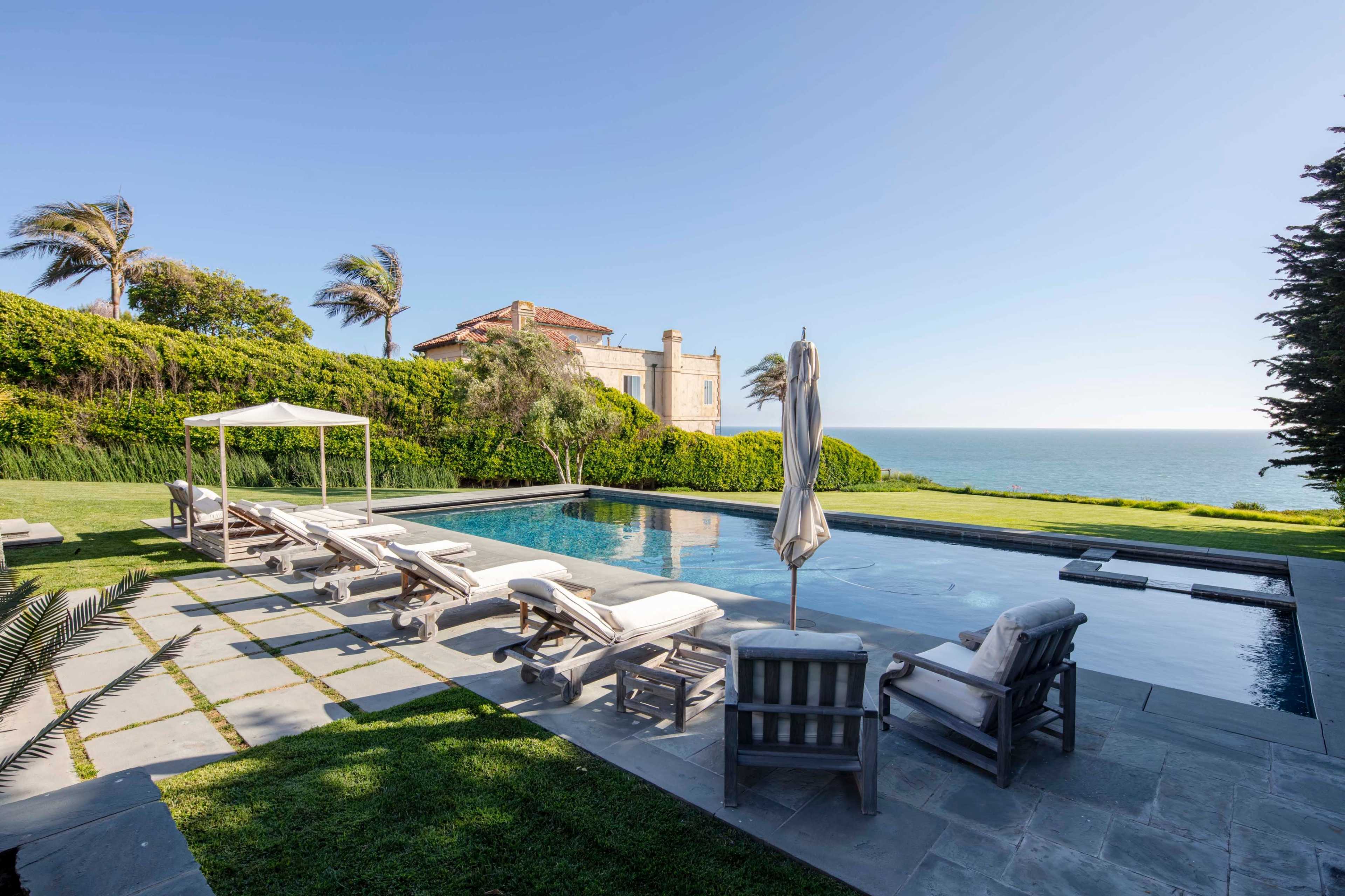 A poolside area with lounge chairs and a canopy overlooking the ocean, surrounded by grass and palm trees.
