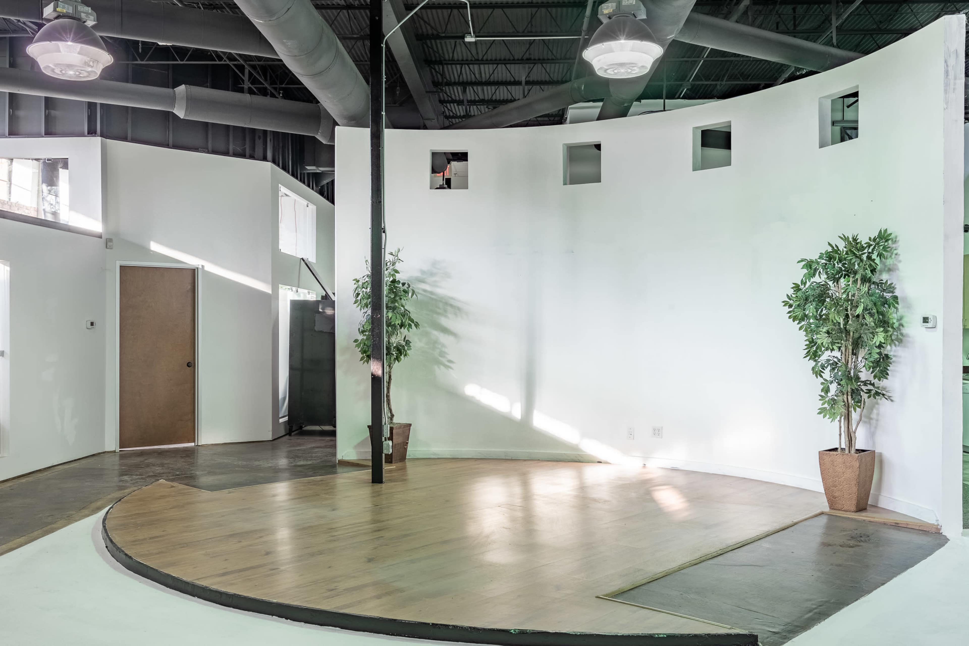 The image shows a spacious, curved interior room with a wooden floor, white walls, and two potted plants.