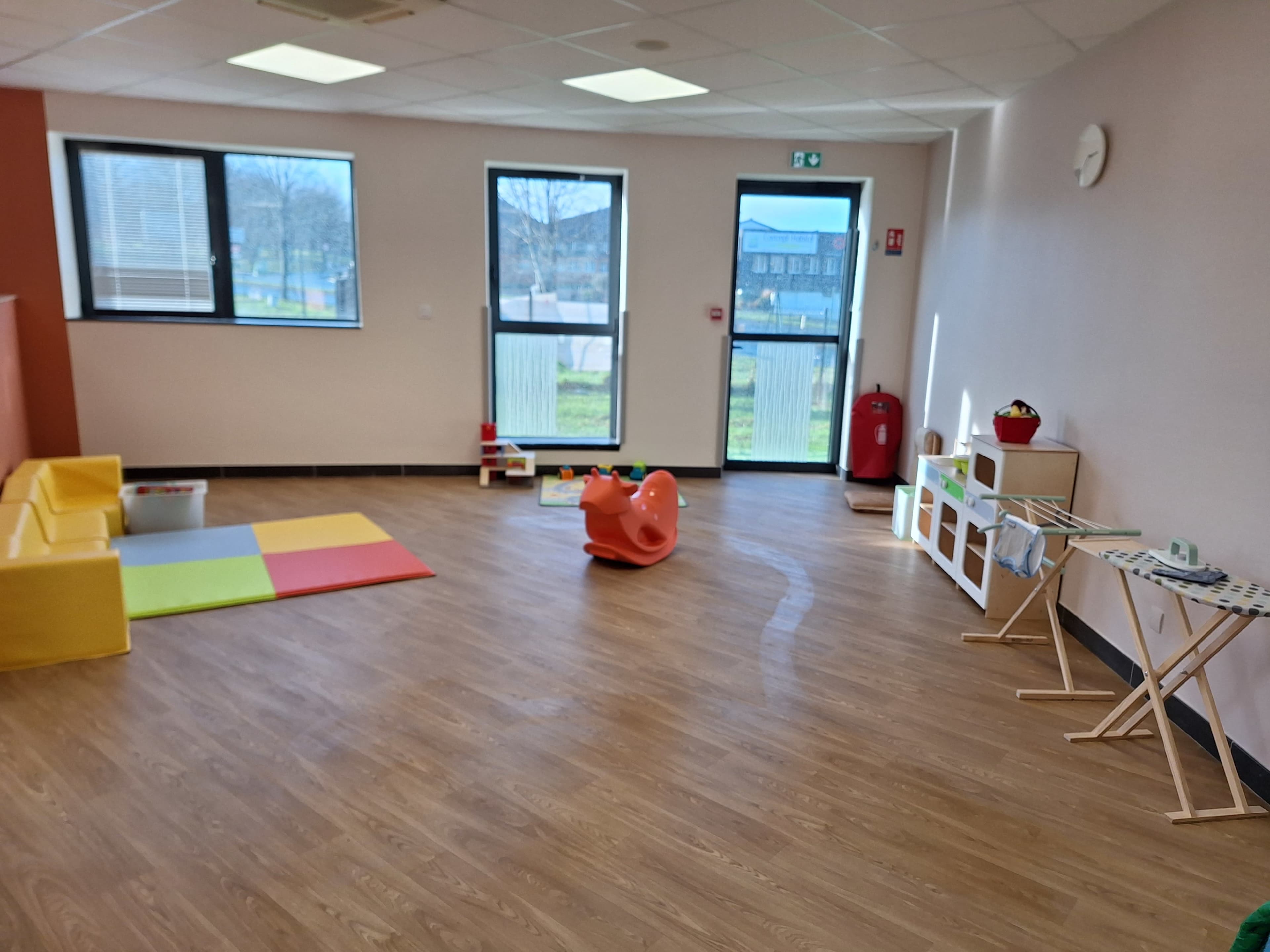 The image shows a bright, spacious playroom with colorful mats, a small slide, and various children’s toys arranged neatly.