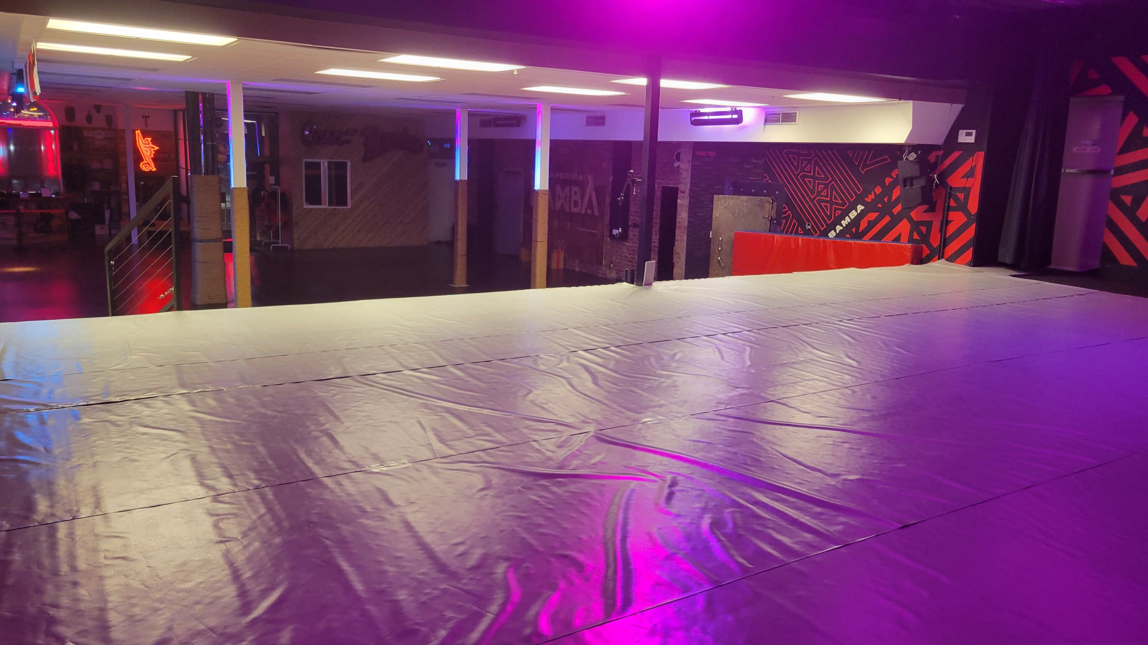 The image shows a spacious indoor venue with a raised platform covered in a smooth surface, illuminated by purple lighting and featuring colorful wall designs in the background.