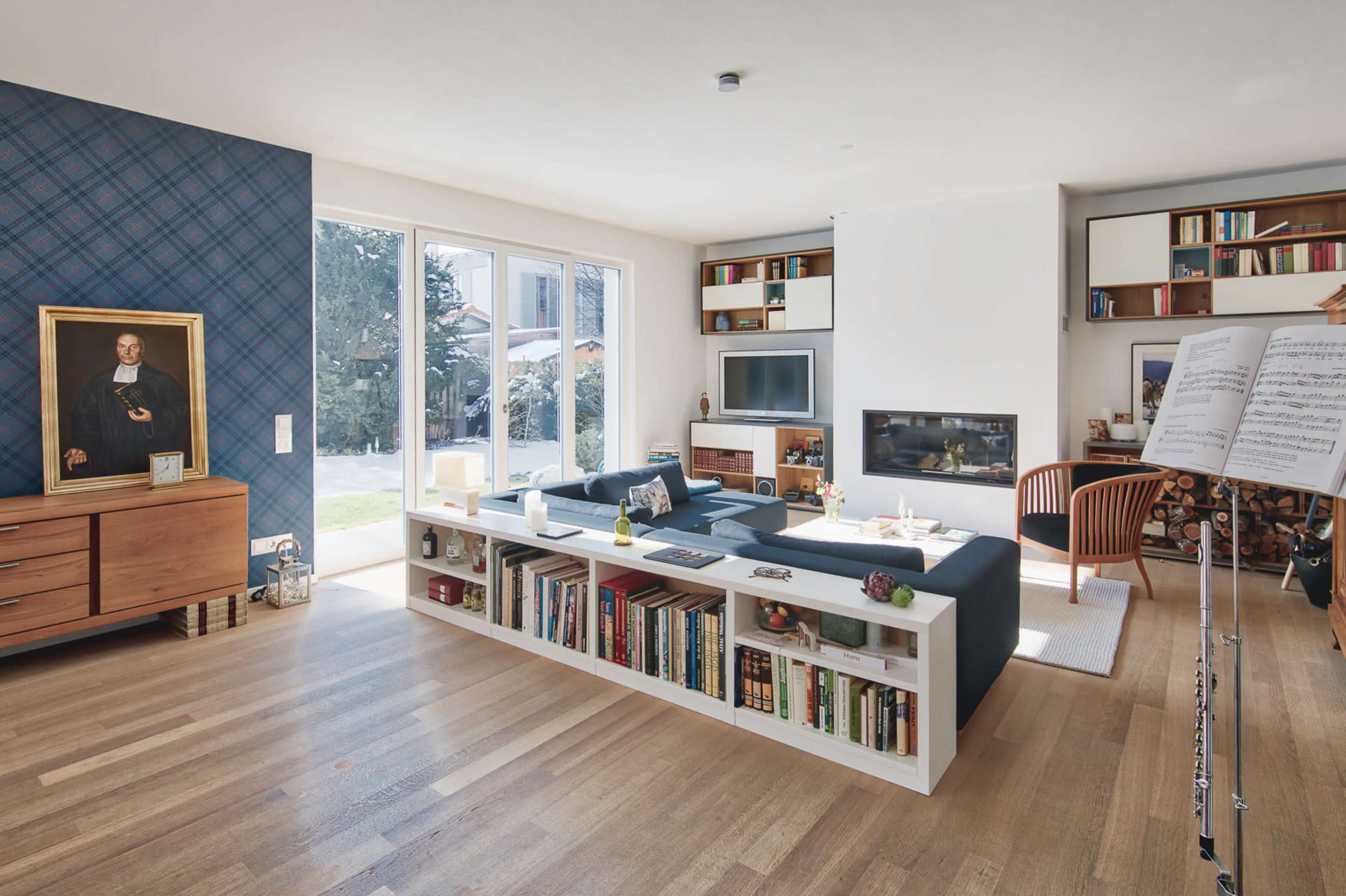 The image shows a modern living room featuring a large bookshelf as a central piece, a flat-screen TV, and large windows overlooking a garden.