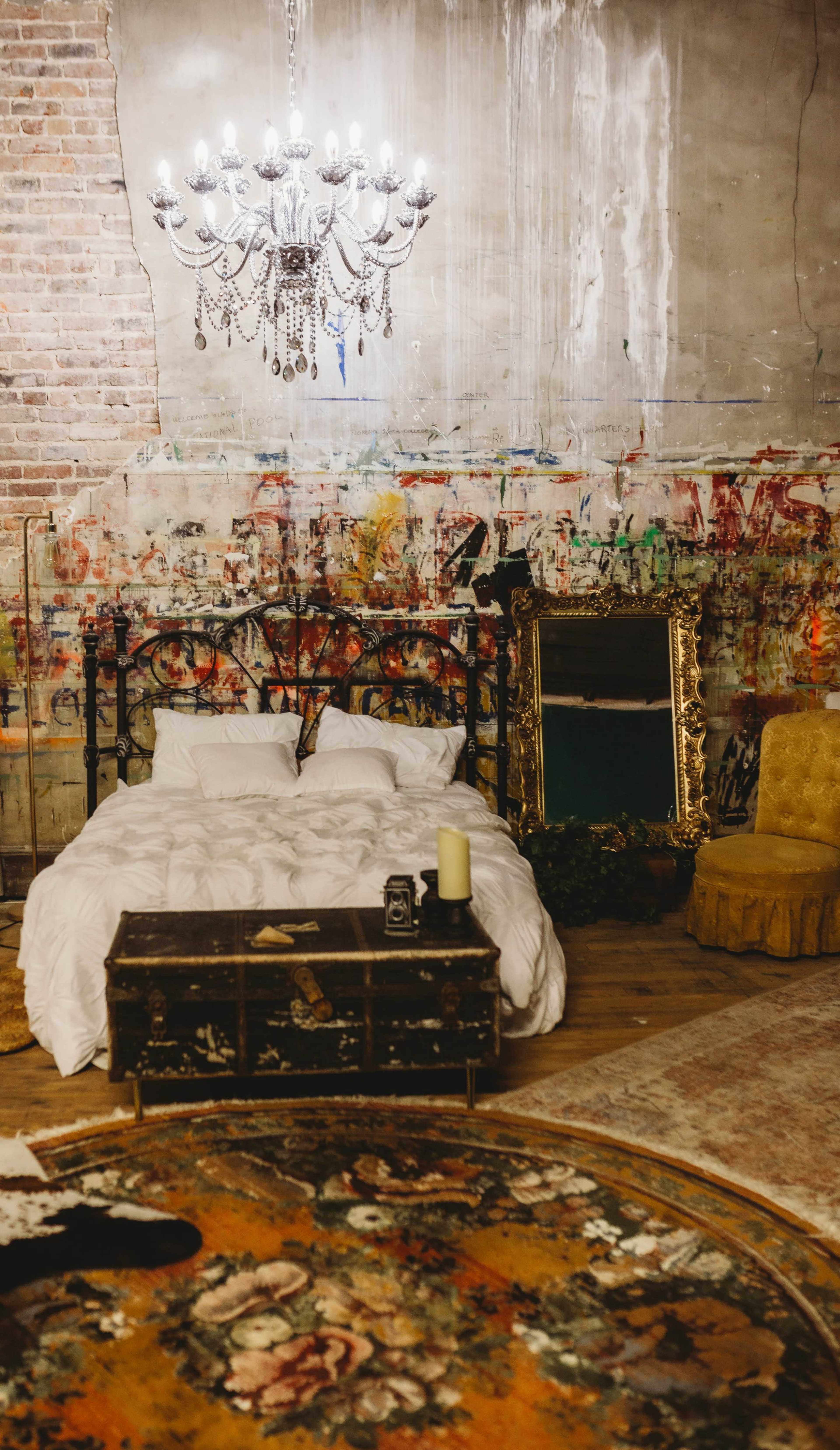 A bedroom features a wrought-iron bed with white bedding, a vintage chandelier, a distressed brick wall covered in colorful paint, and a gold-framed mirror beside a decorative yellow chair.