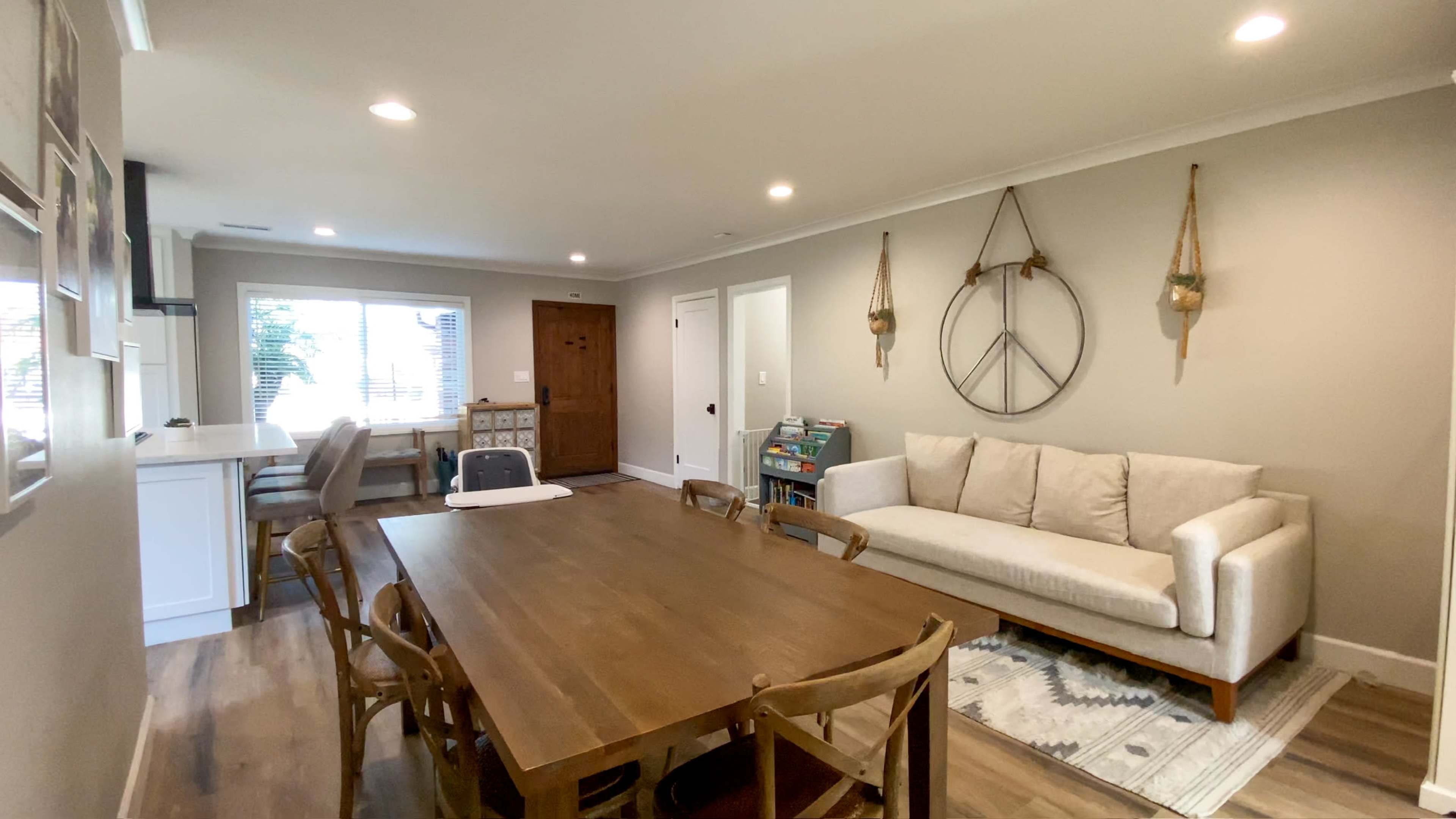 The image shows a well-lit dining area with a wooden table, chairs, and a cozy couch alongside a window and a door leading outside.