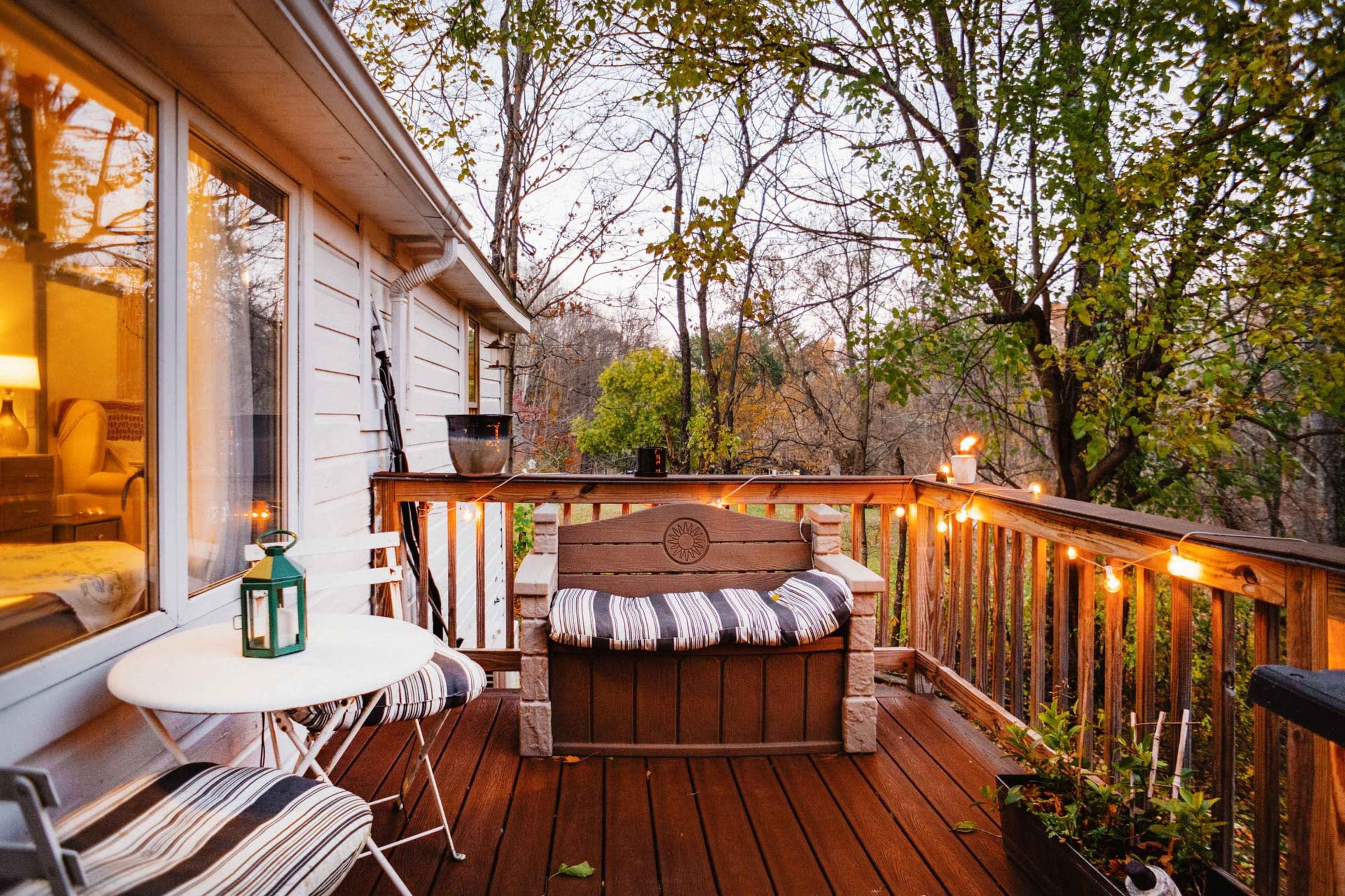 A wooden deck features a seating area with a striped cushion, a small table, and string lights surrounded by trees.
