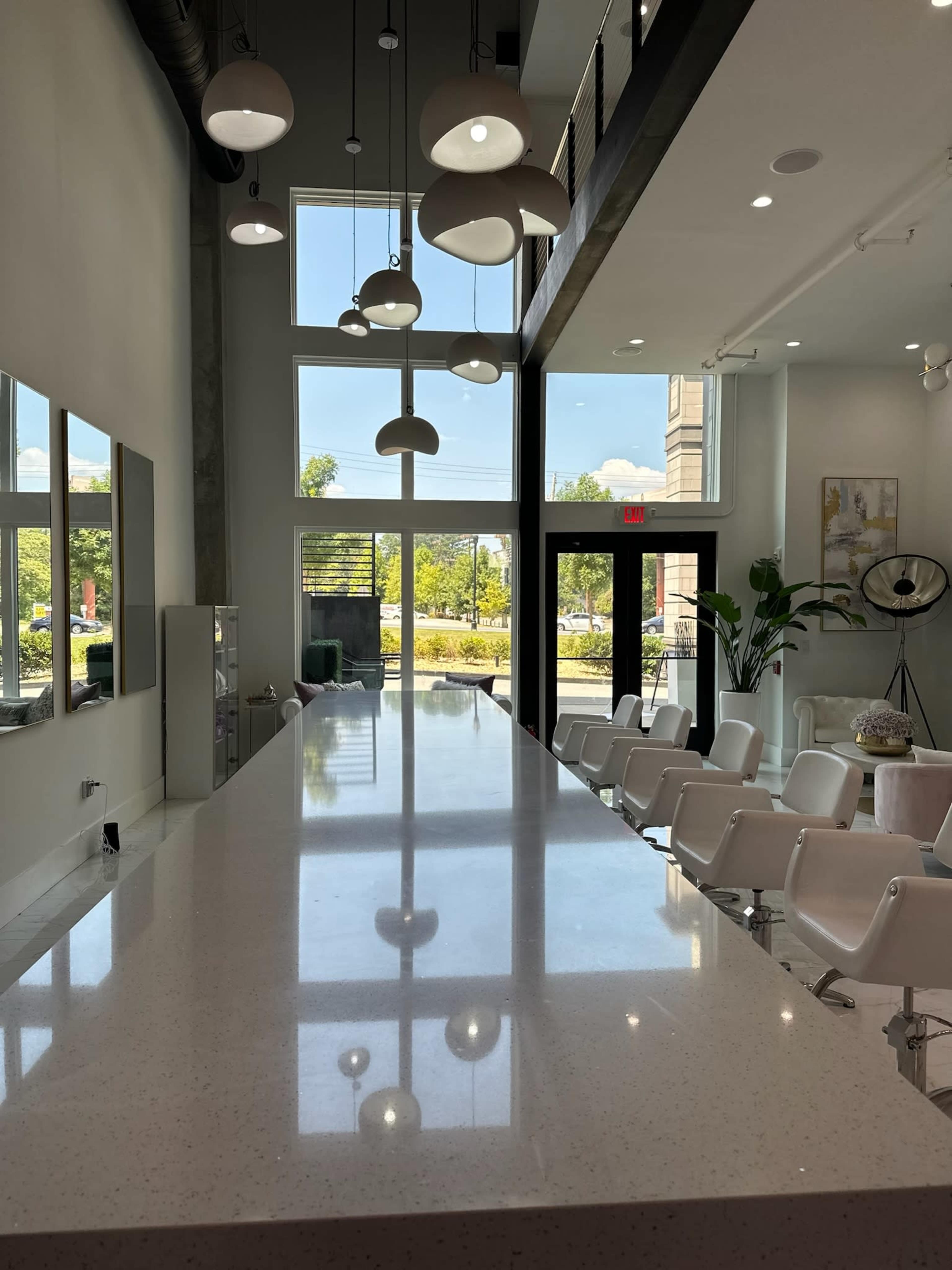 The image shows a modern indoor space with a long countertop, large windows, and pendant lighting, featuring white chairs arranged for seating.