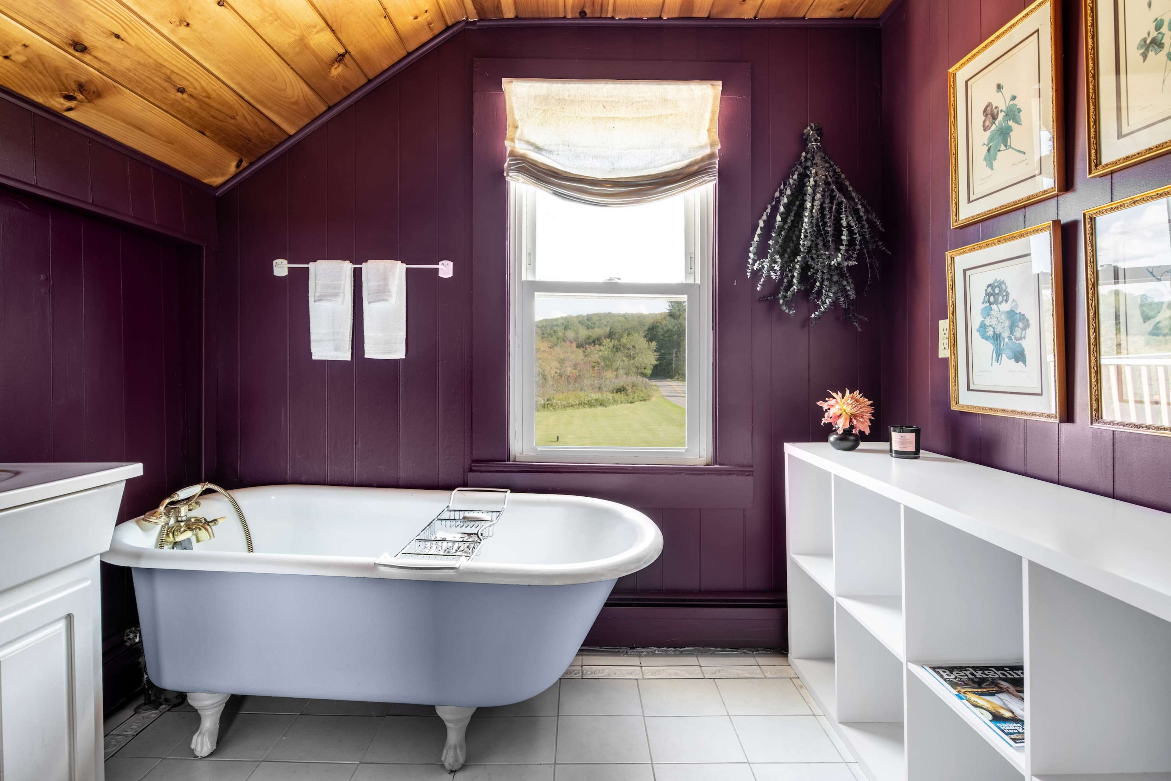 A bathroom with a vintage clawfoot bathtub, purple walls, a window providing natural light, and a small shelf with decor.