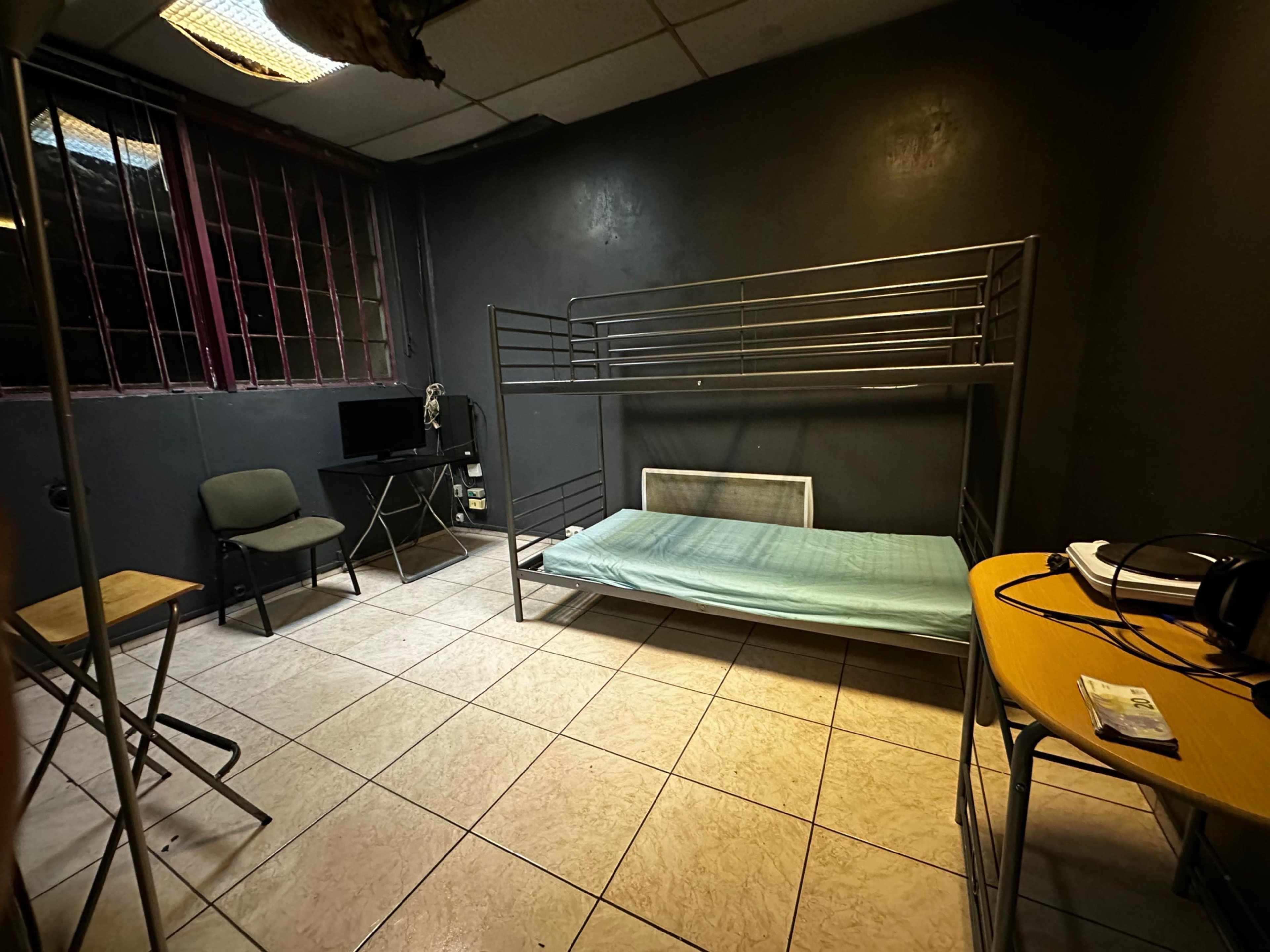 The image shows a sparsely furnished room with a bunk bed, a small desk and chair, a laptop on a stand, and a window with barred grilles.