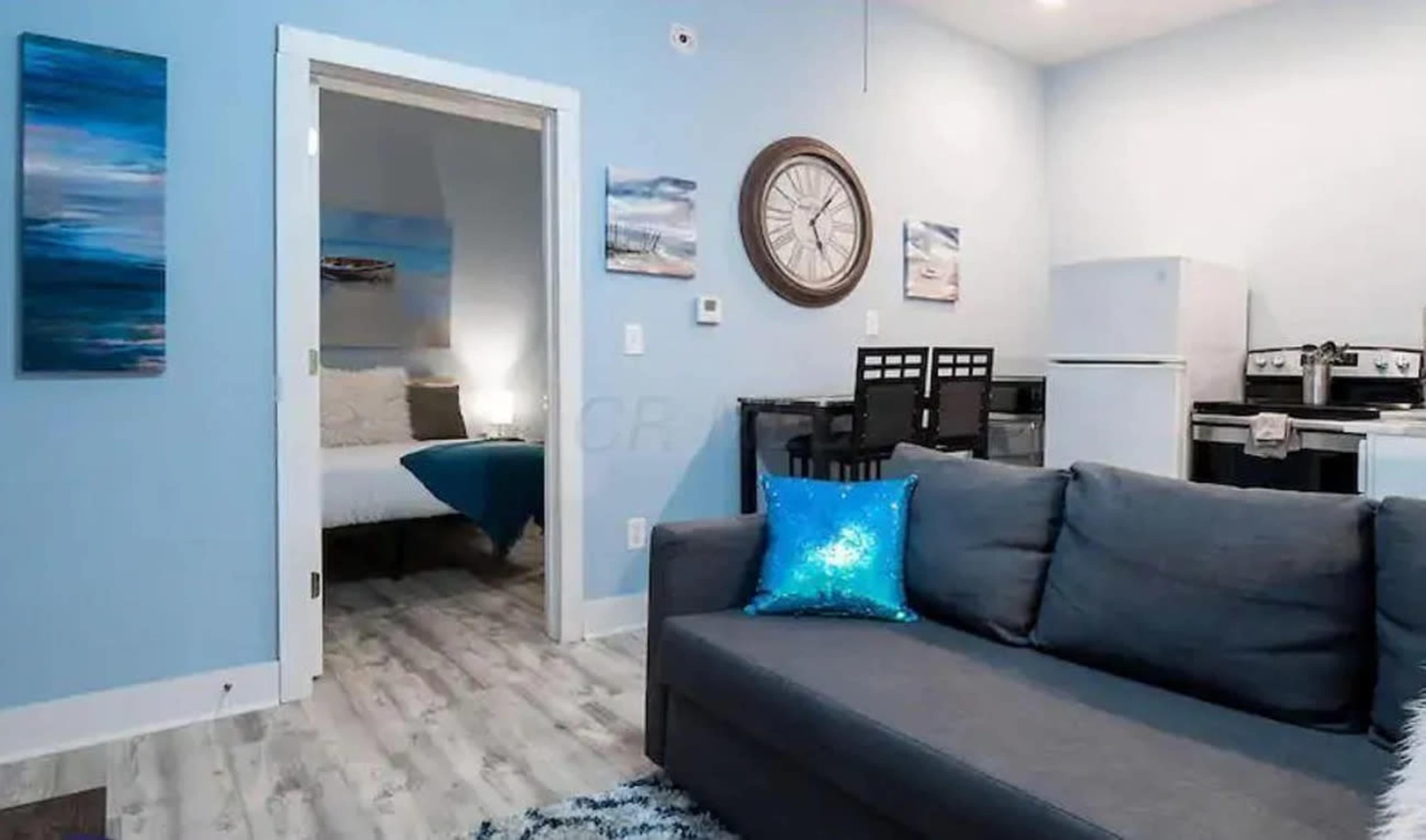 The image shows a living area with a gray sofa, a blue decorative pillow, and an open doorway leading to a bedroom, with a kitchen visible in the background.