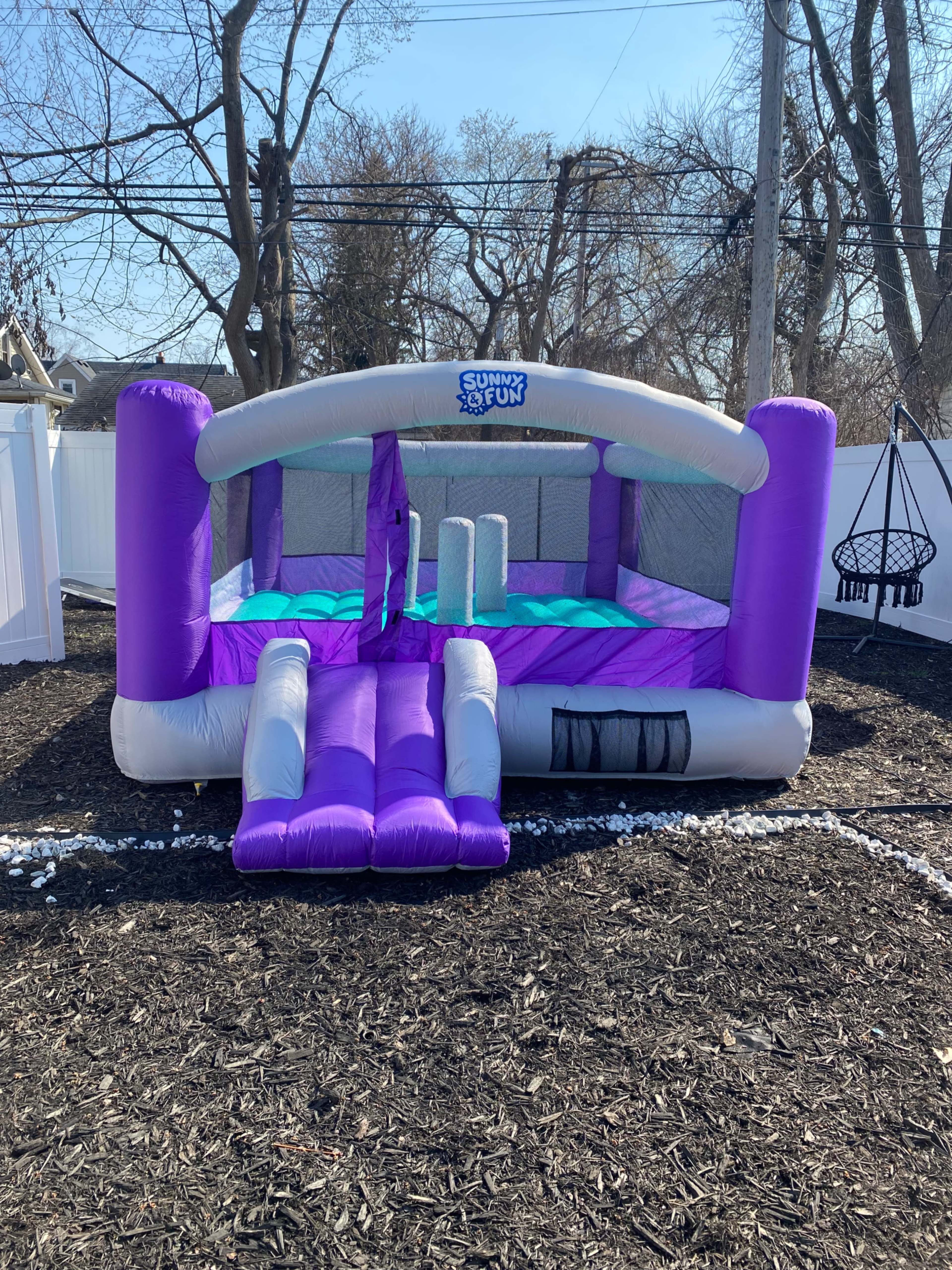 The image shows a purple and gray inflatable bounce house with a slide, set up in a backyard with dark mulch and a white fence in the background.