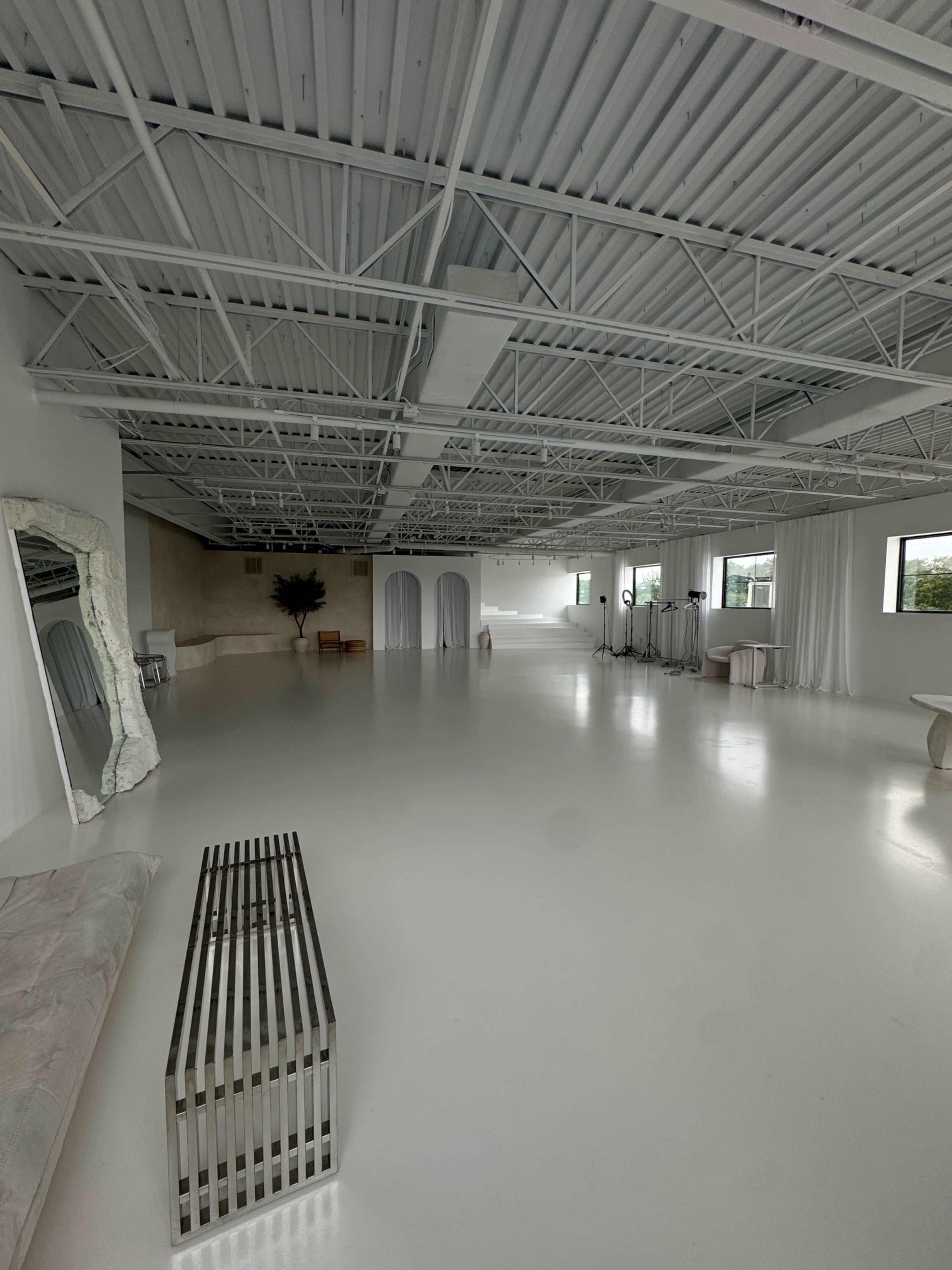 The image shows a spacious, minimalistic interior with a glossy white floor, large windows, and a few pieces of furniture including a mirror and a striped table.