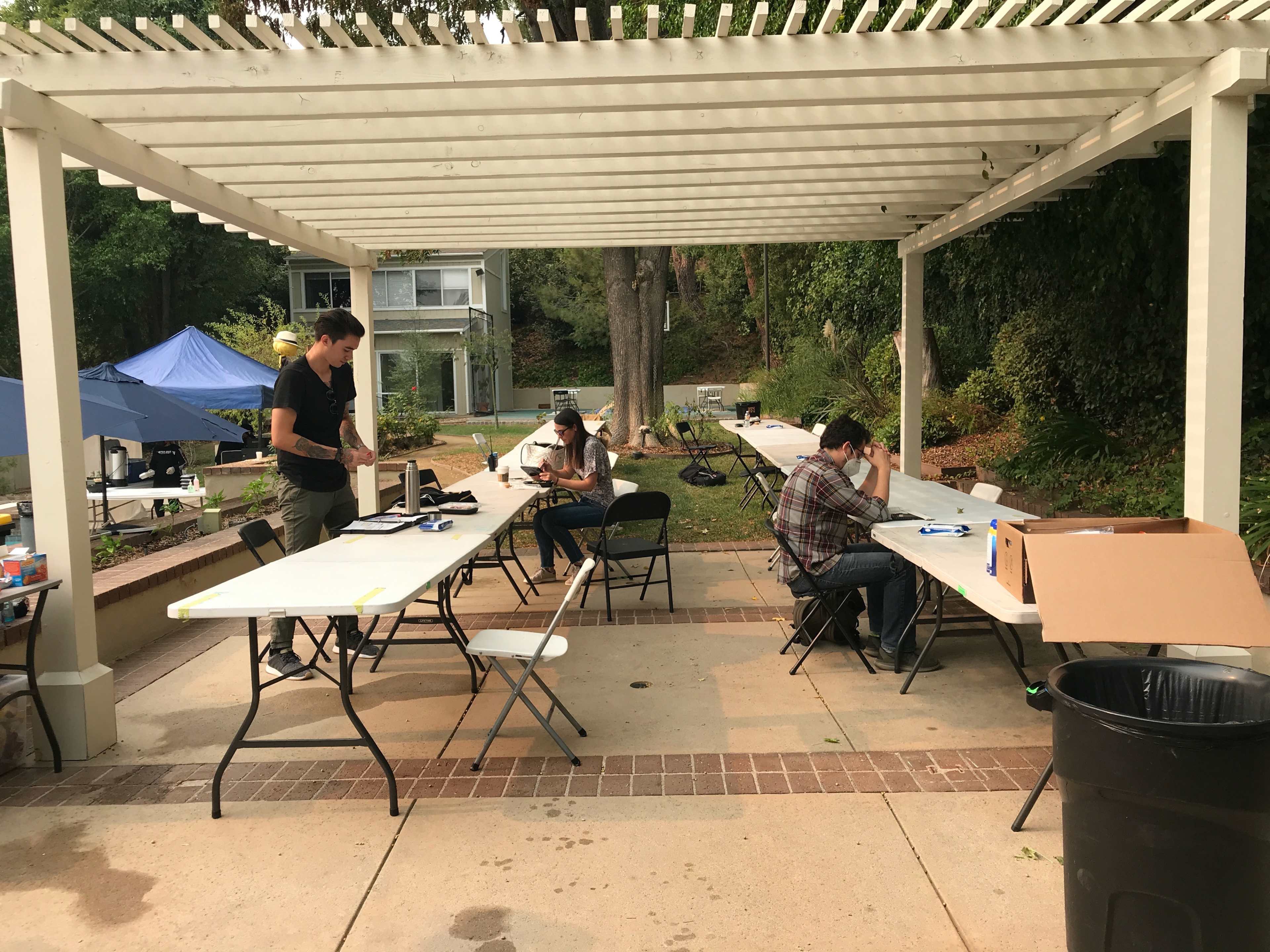 A group of individuals is working at tables under a pergola in an outdoor setting.