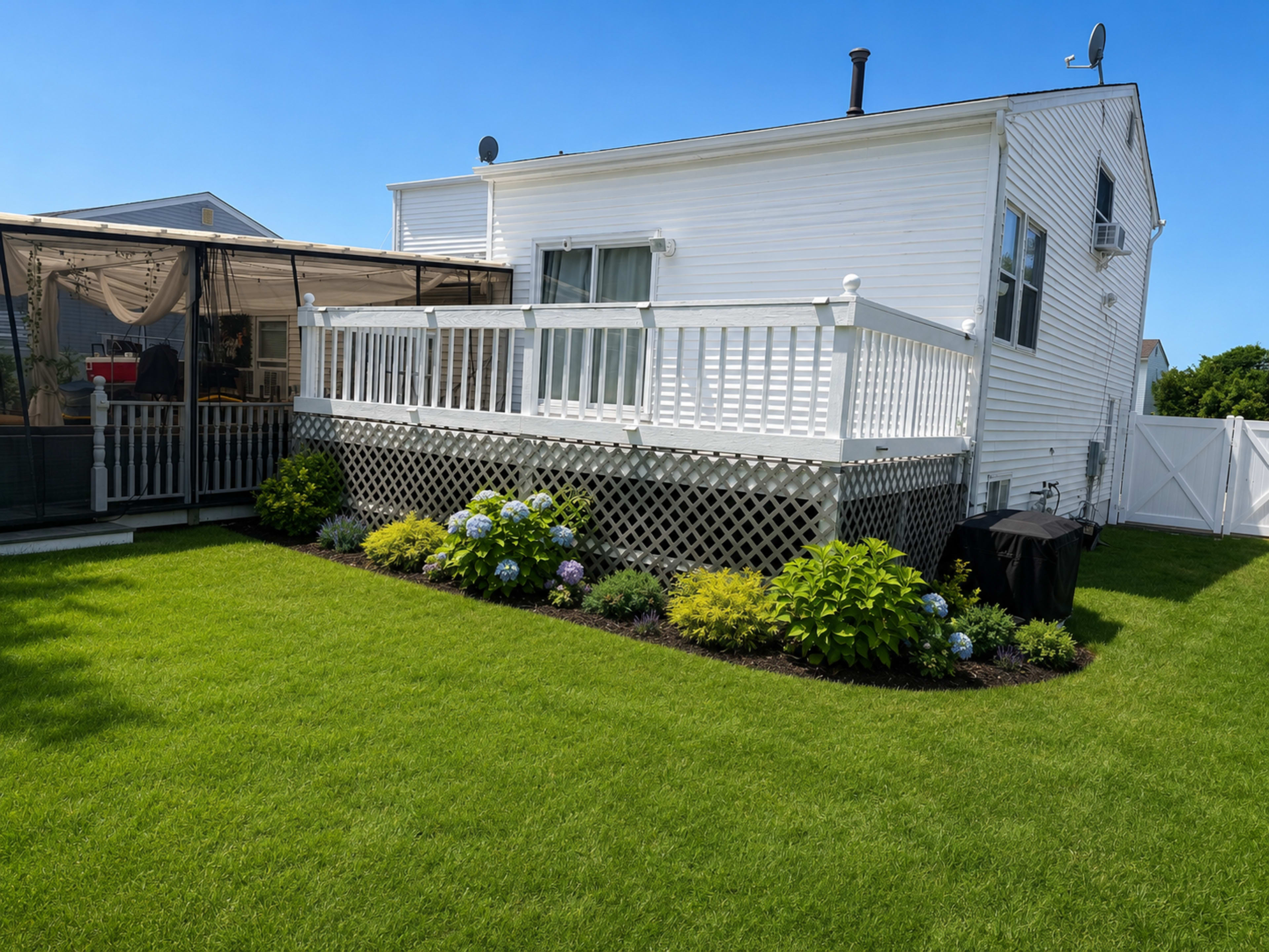 A white house with a deck is surrounded by well-maintained grass and landscaped flower beds featuring hydrangeas.