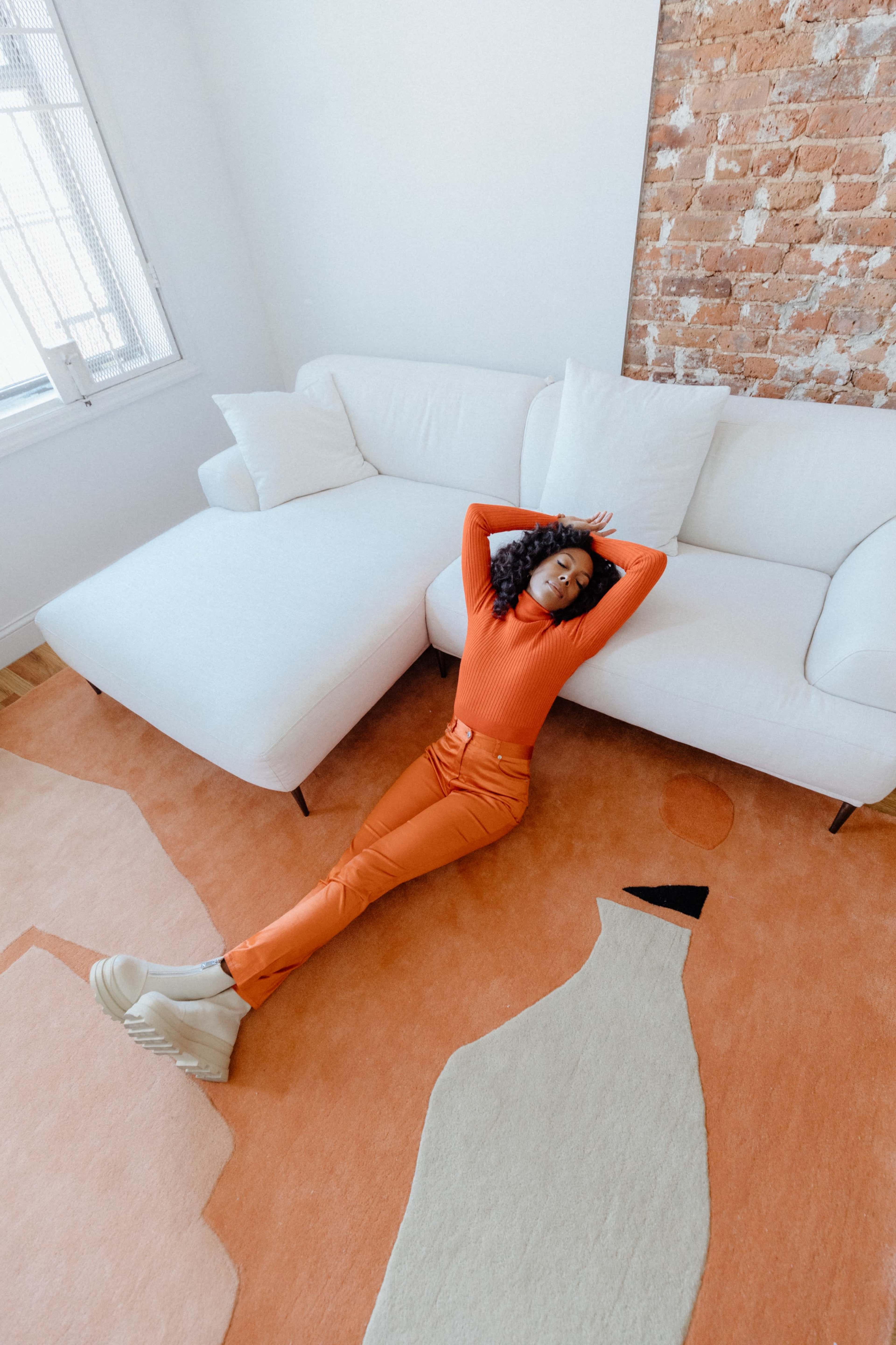 A person in an orange outfit relaxes on a carpet while lying on the floor beside a white sectional sofa in a bright room.