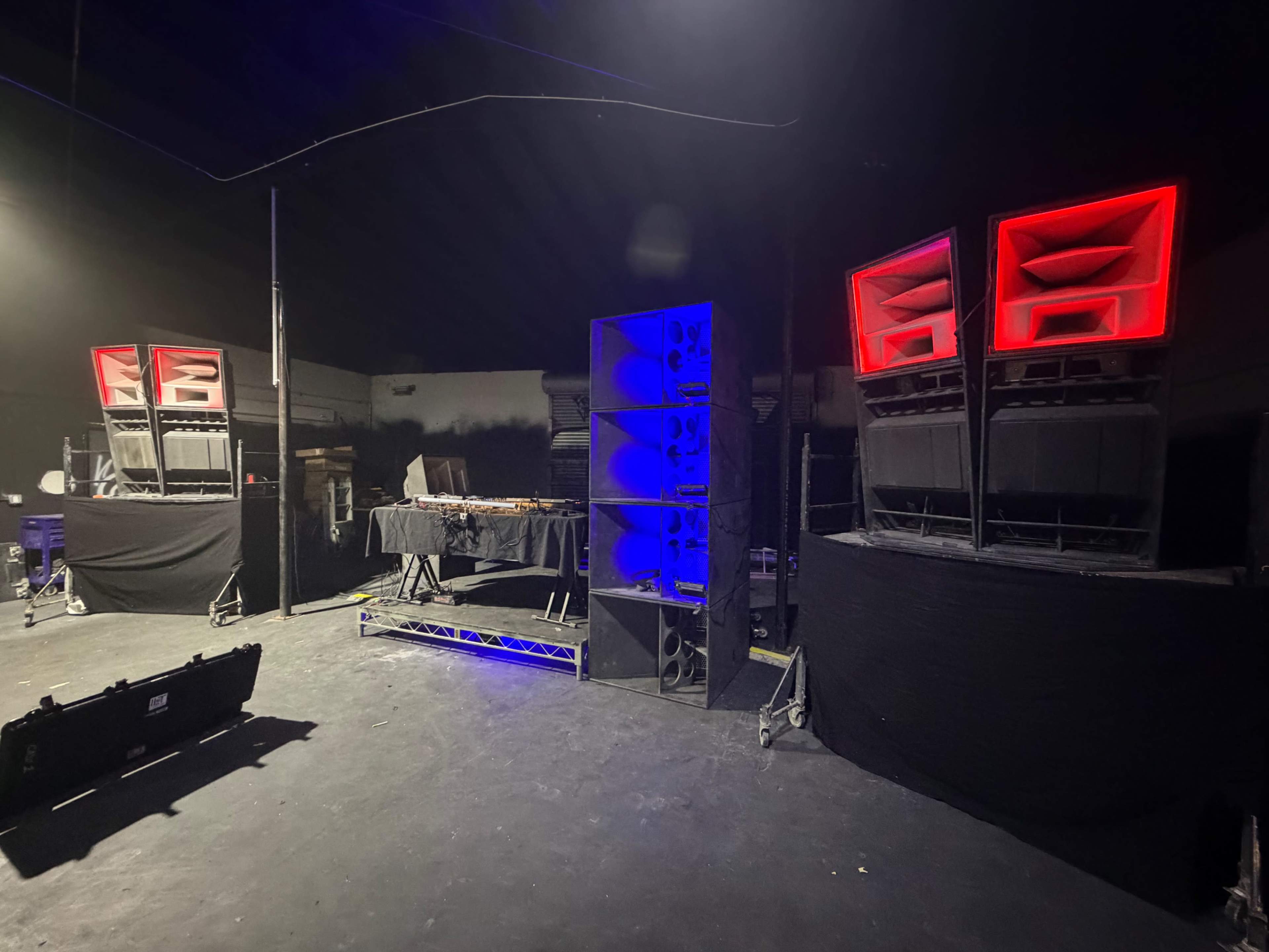The image shows a dimly lit music venue with large sound speakers arranged on either side of a DJ booth.