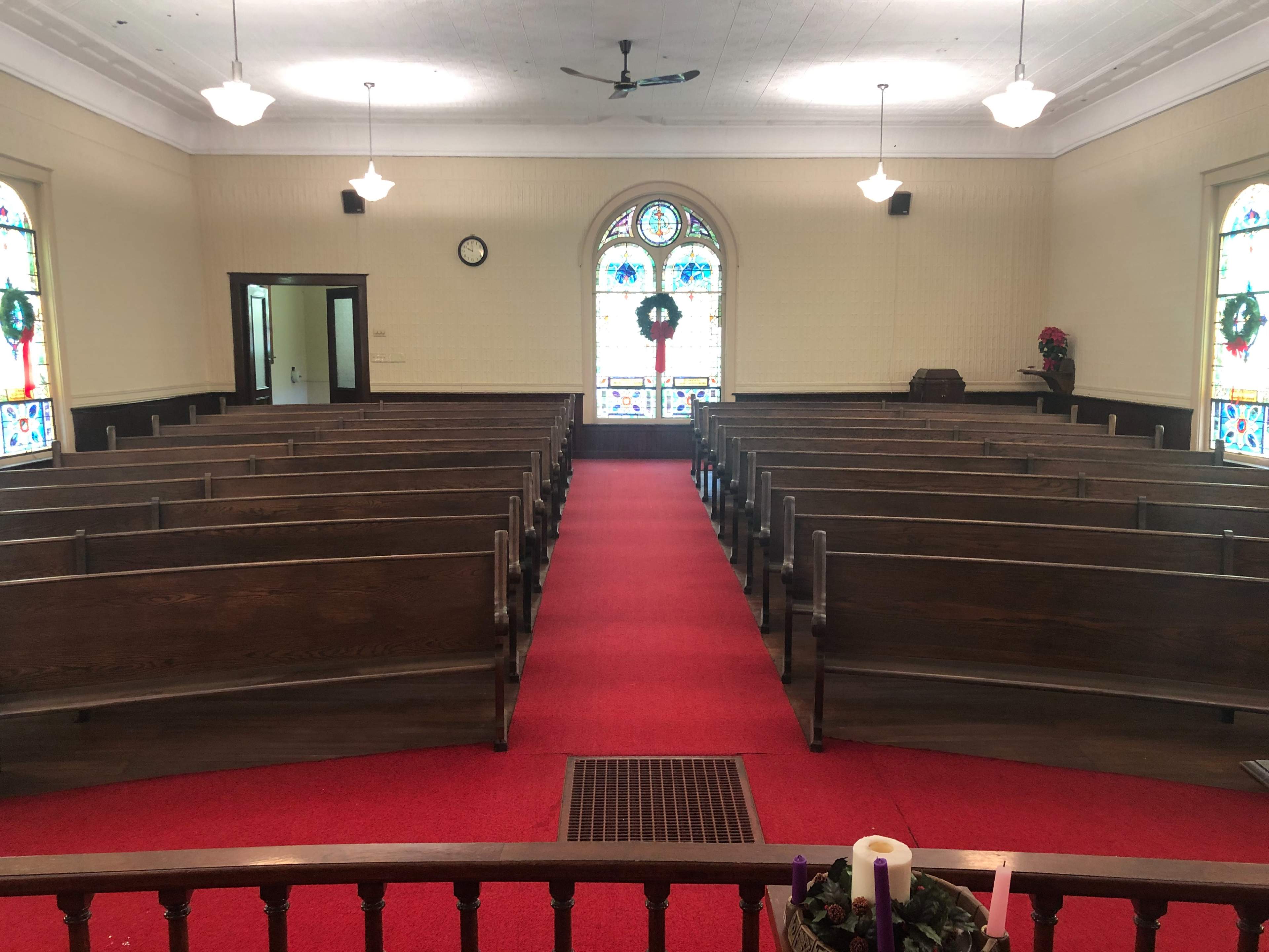A church interior with wooden pews arranged in rows on red carpet, facing a stained glass window at the front.