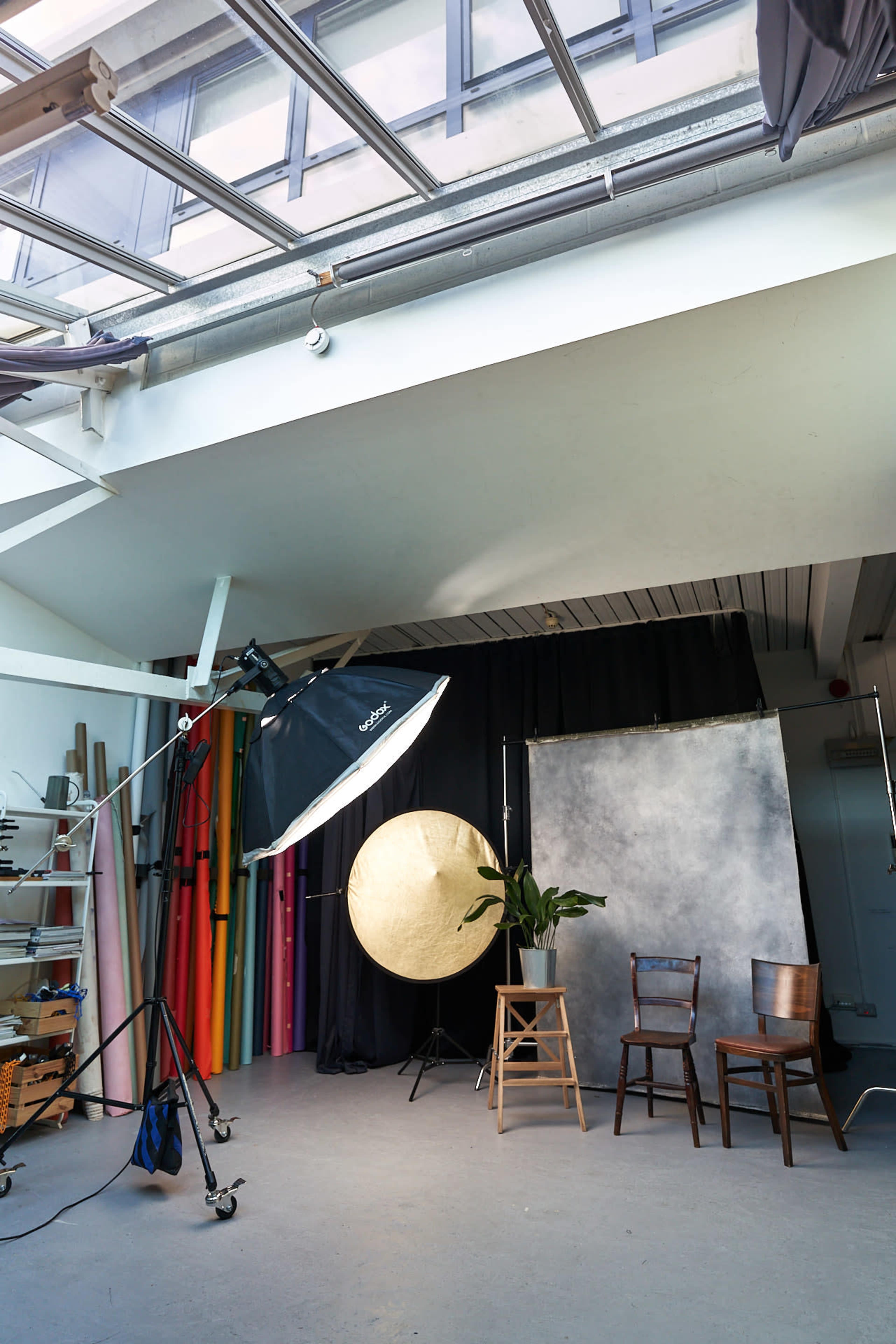 The image shows a photography studio setup with a large softbox light, a circular reflector, a backdrop, and two wooden chairs.