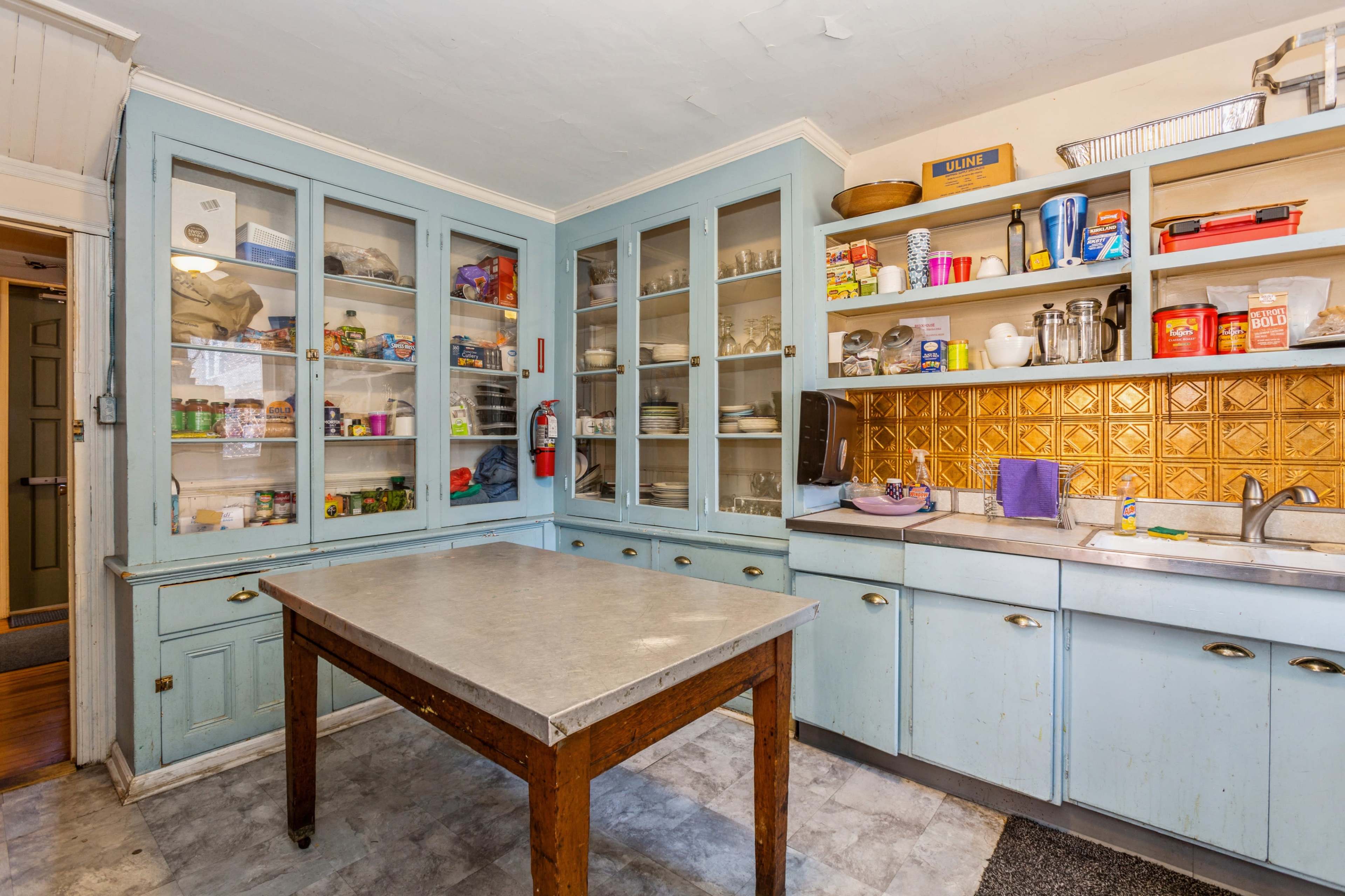 A kitchen with light blue cabinets, a large table in the center, and shelves filled with various items and utensils.