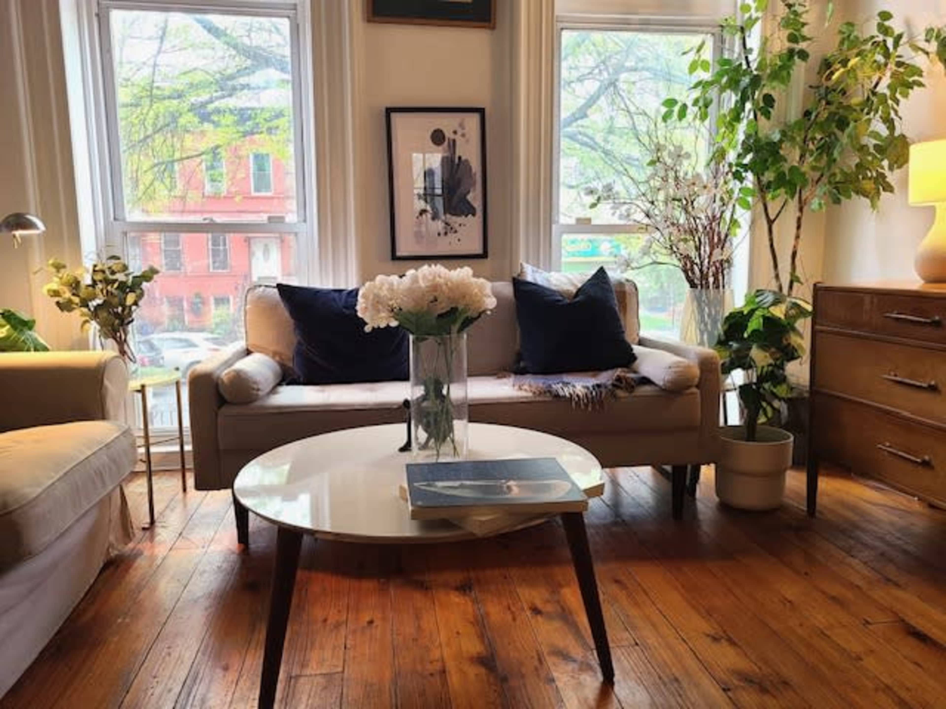 A cozy living room with a white sofa, a round coffee table, and large windows providing natural light, surrounded by indoor plants and decorative items.