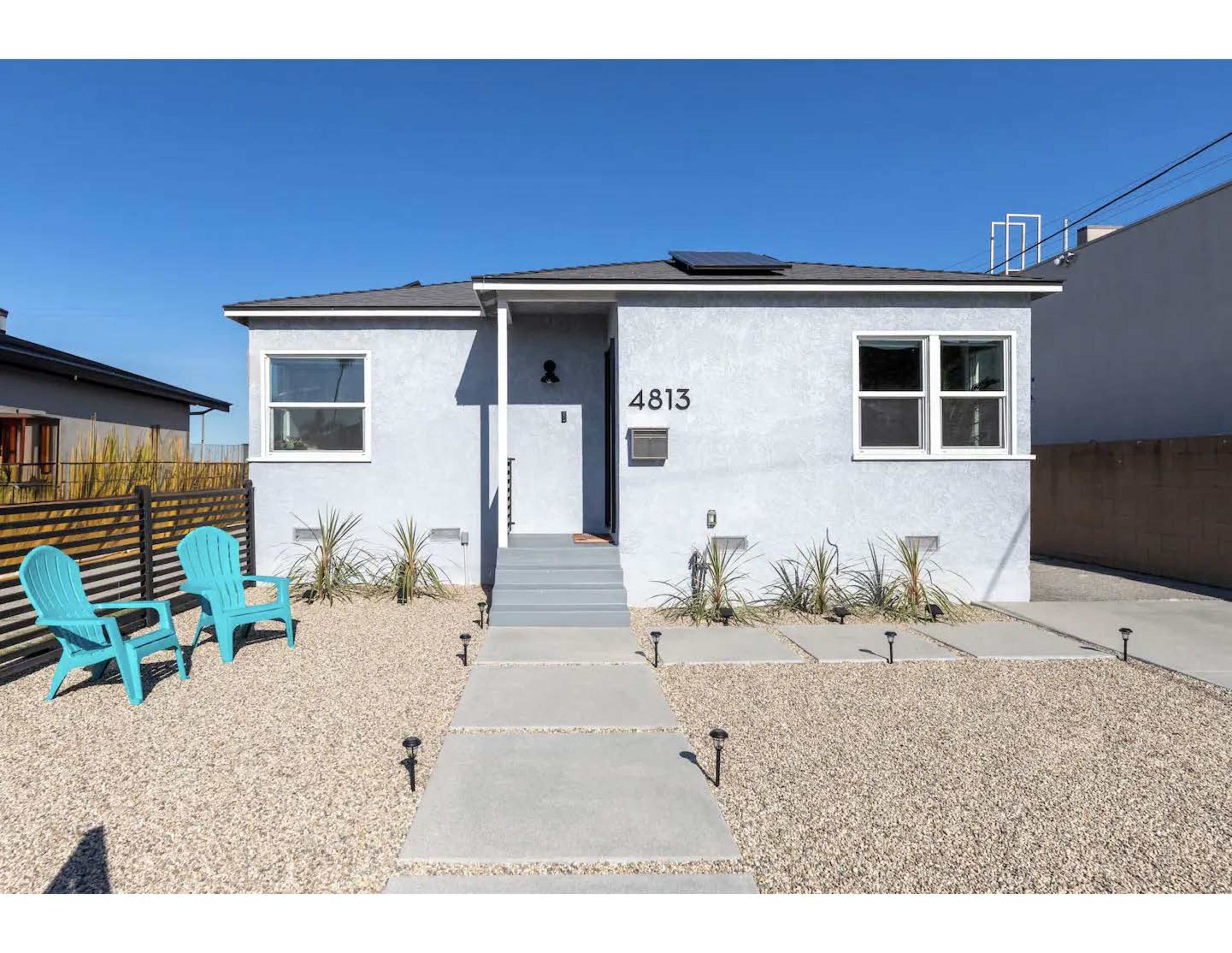 The image shows a single-story house with a light-colored exterior, a small front staircase, and two turquoise chairs positioned on the gravel front yard.