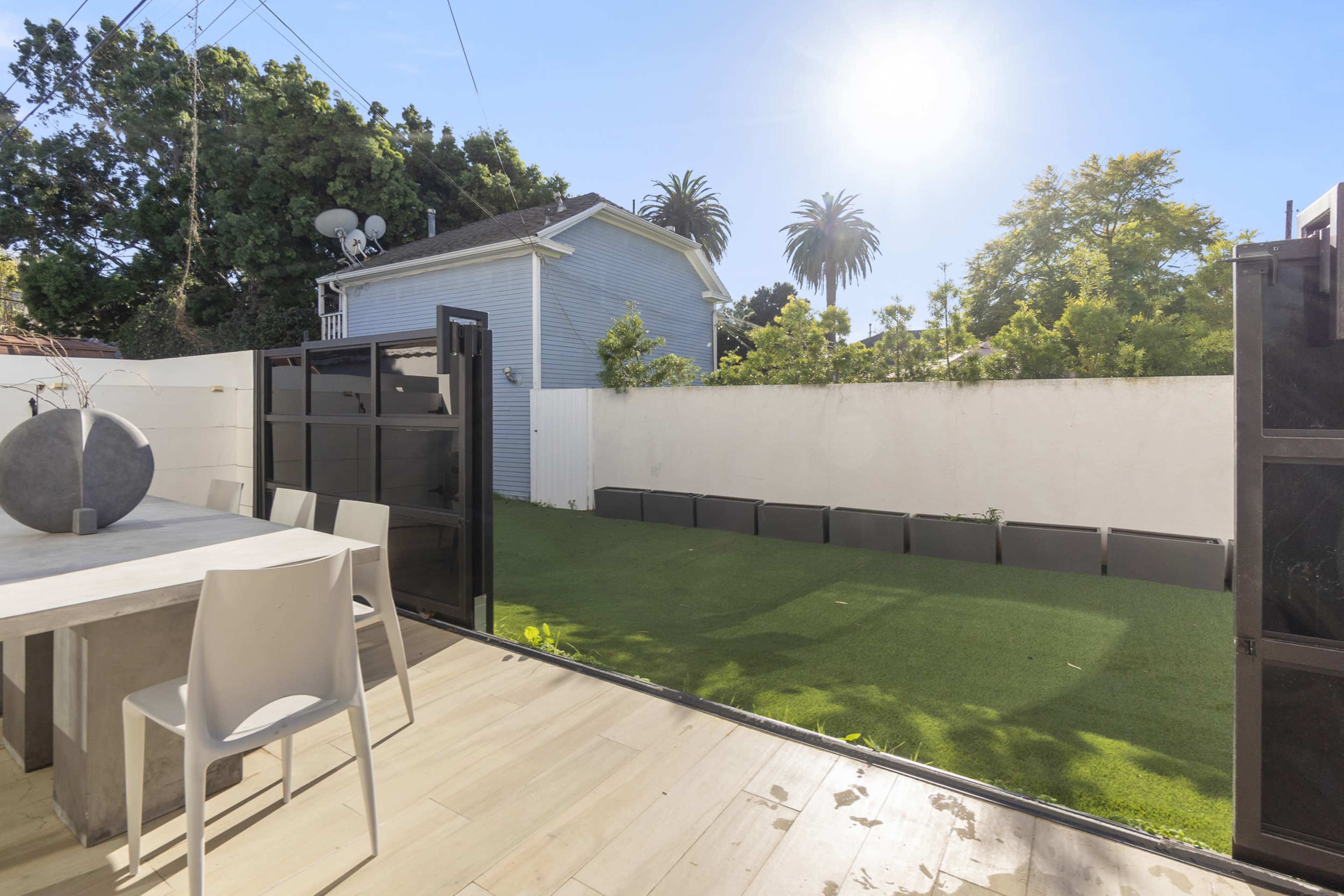 A patio with a modern table and chairs opens up to a green lawn surrounded by white walls and trees under a sunny sky.
