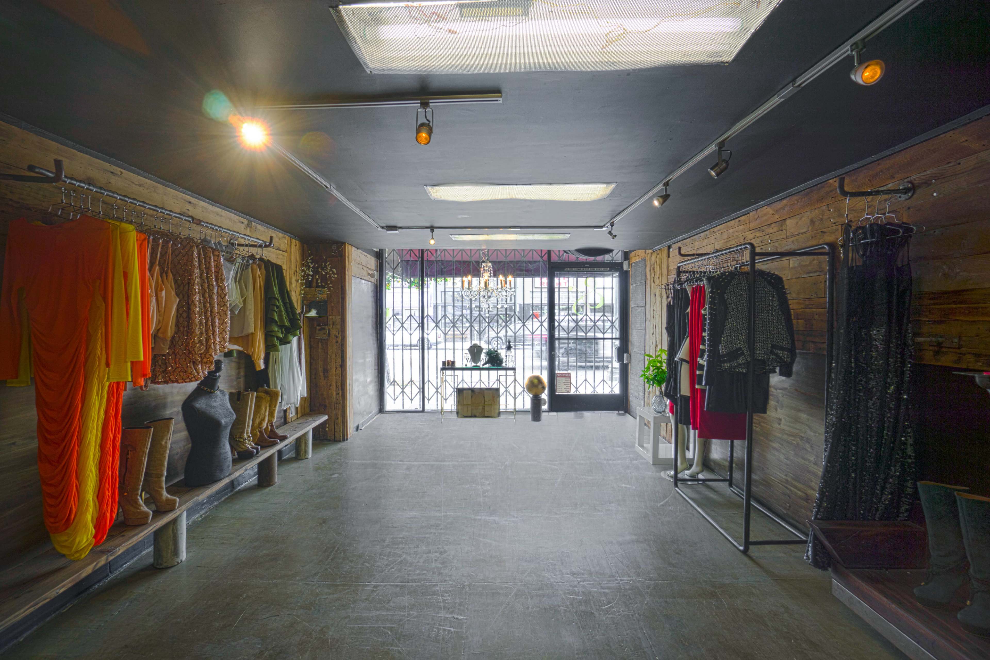 The interior of a clothing boutique features clothing racks with various garments, wooden accents, and a large window with barred security.