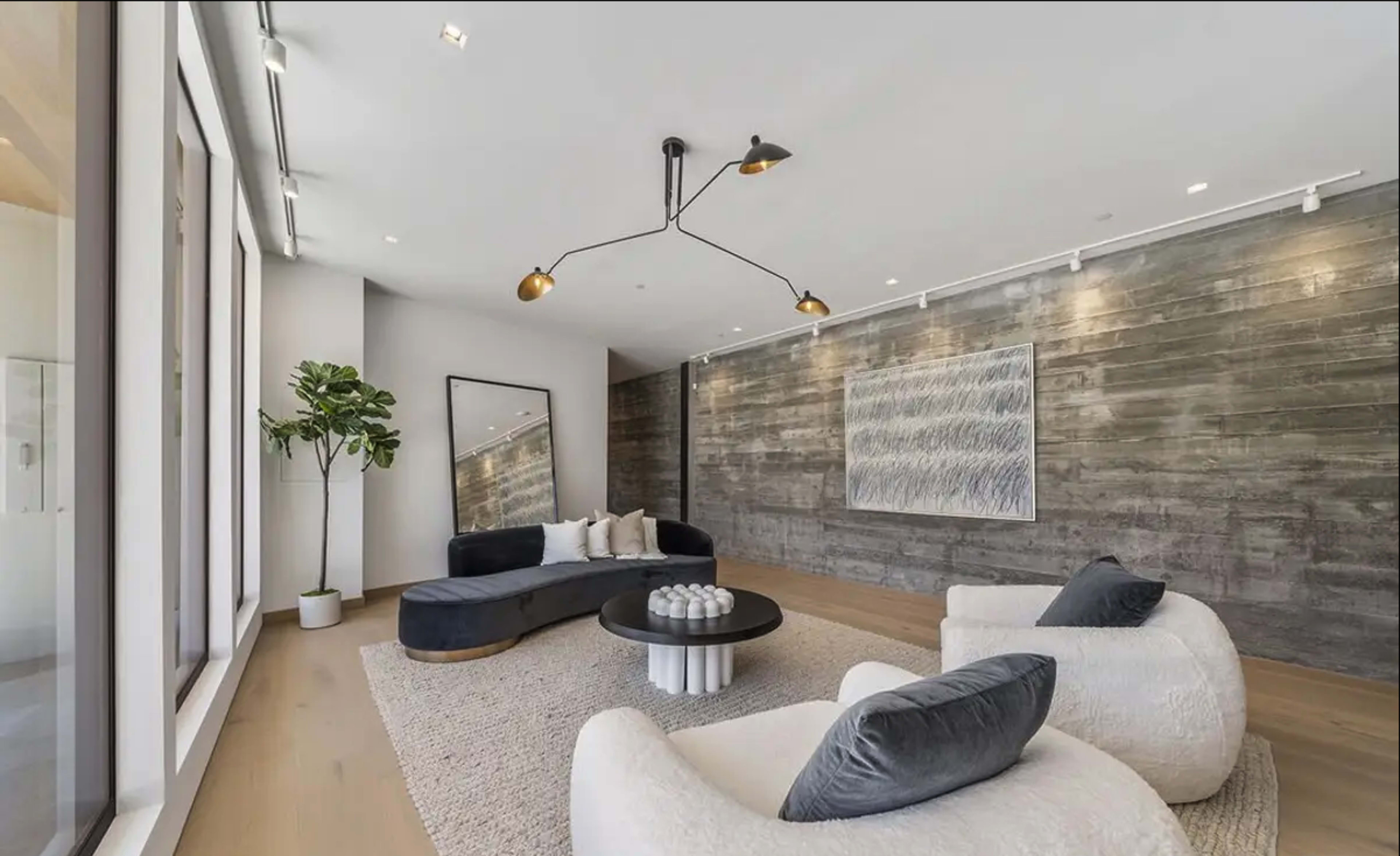 A modern living room featuring a curved dark sofa, two upholstered chairs, a round coffee table with decorative items, and a large mirror on one wall.
