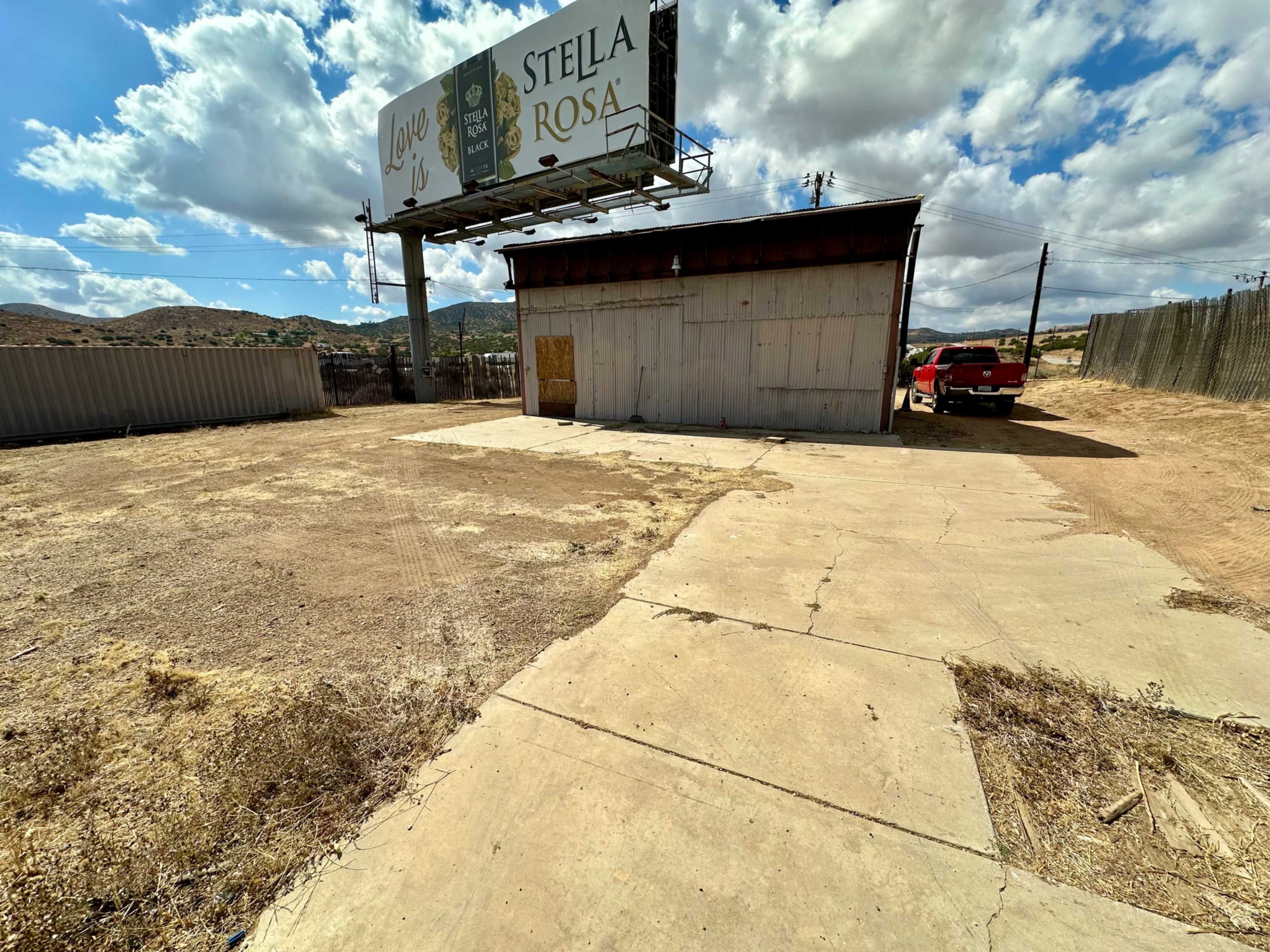 The image shows a vacant lot with a small, boarded-up building and a pickup truck parked nearby, under a cloudy sky with distant hills in the background.