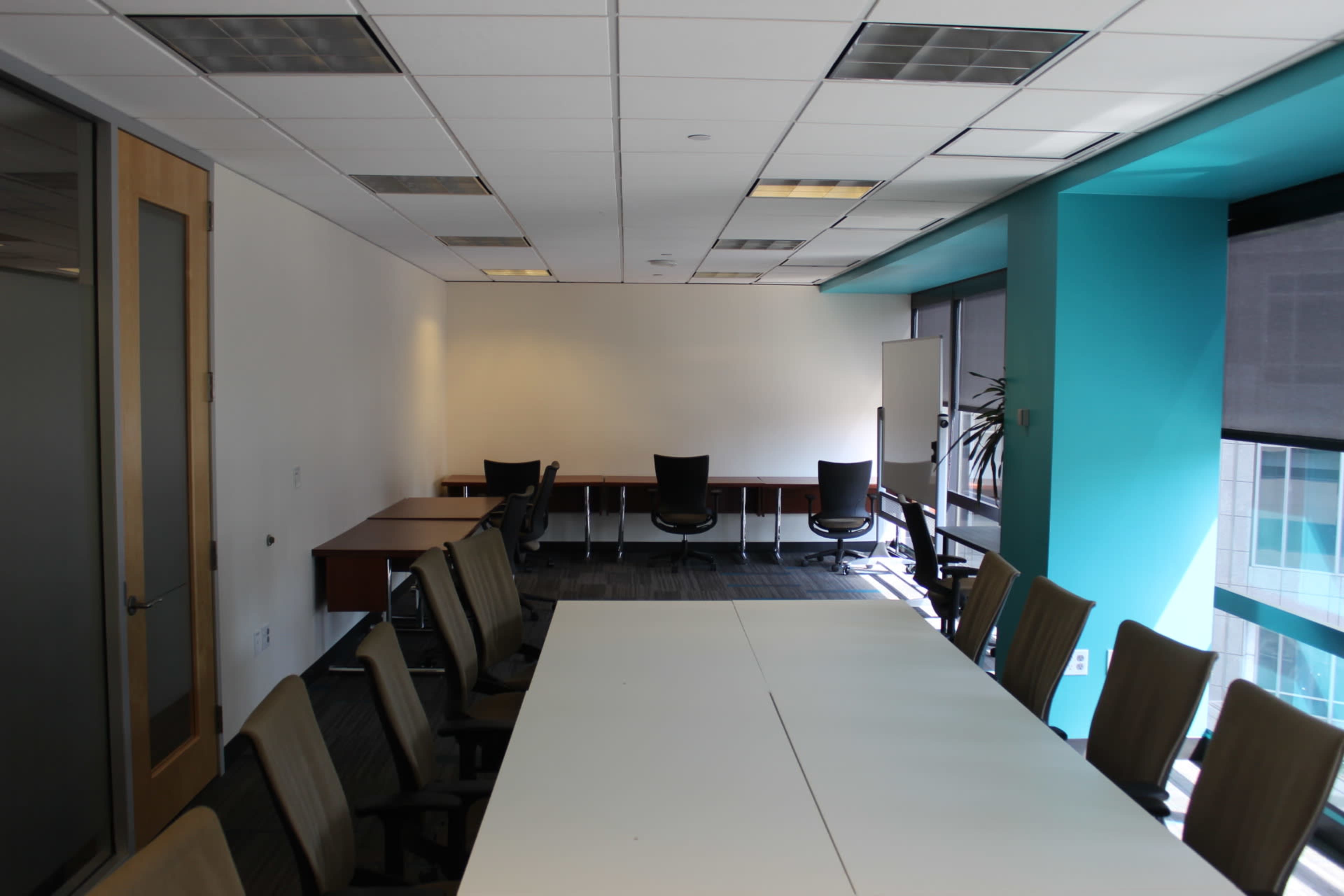 A conference room with a large table in the foreground, surrounded by chairs, and additional seating along the back wall.
