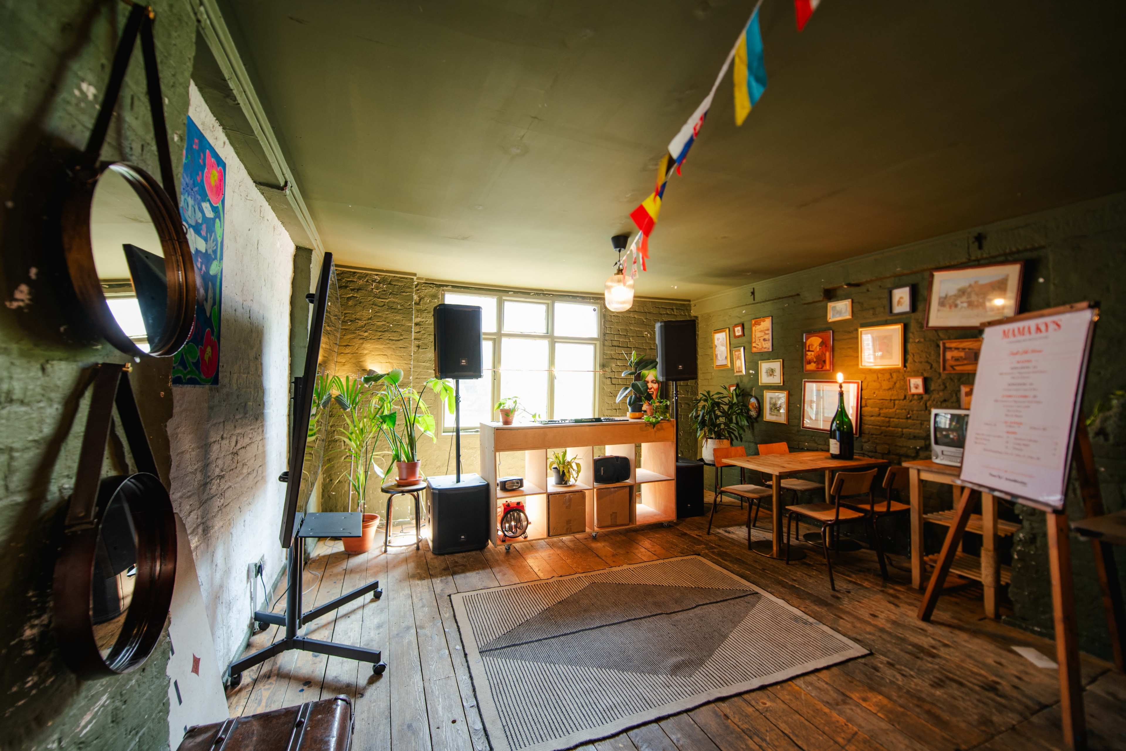 The image shows a cozy indoor space with wooden floors, a small stage, plants, and framed pictures on the walls.