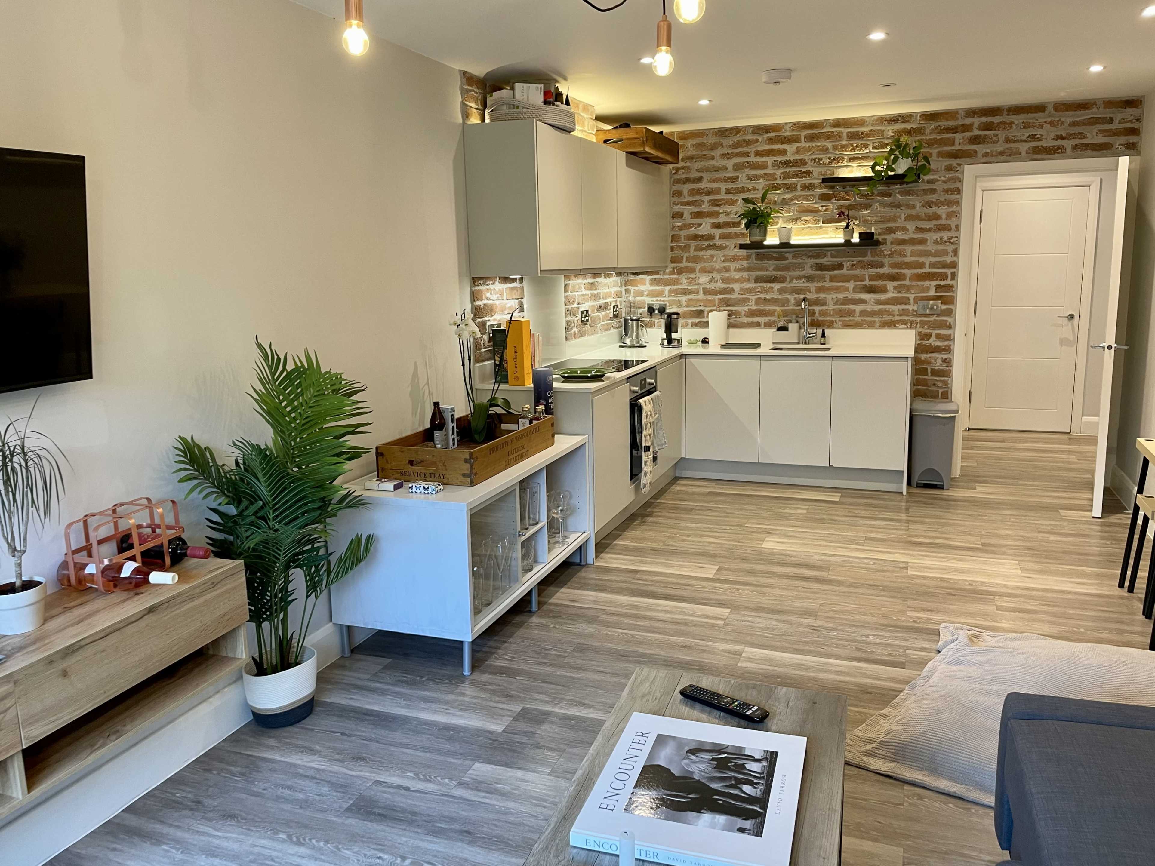 A modern kitchen and living area with a brick accent wall, light wood flooring, and contemporary furnishings including a television, a plant, and a coffee table.