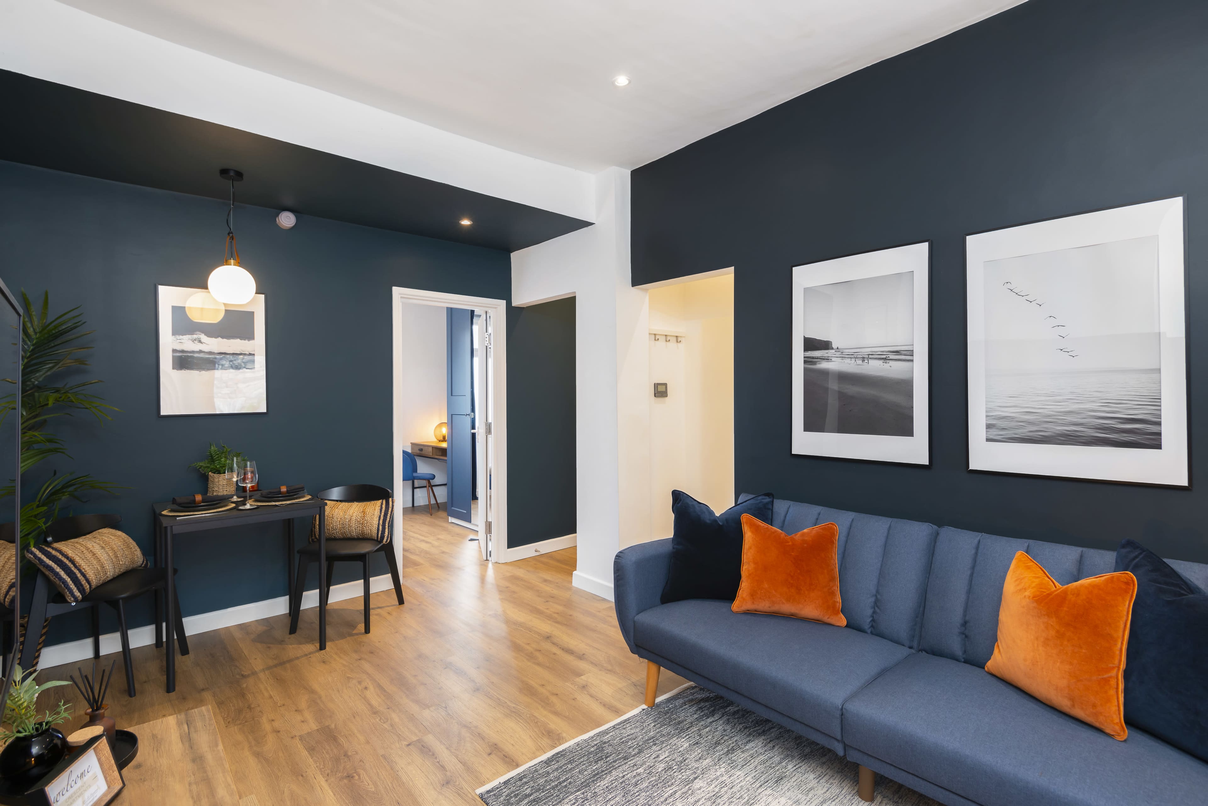 A modern living space featuring a blue couch with orange pillows, a dining area with a table and two chairs, and framed photographs on the dark blue wall.