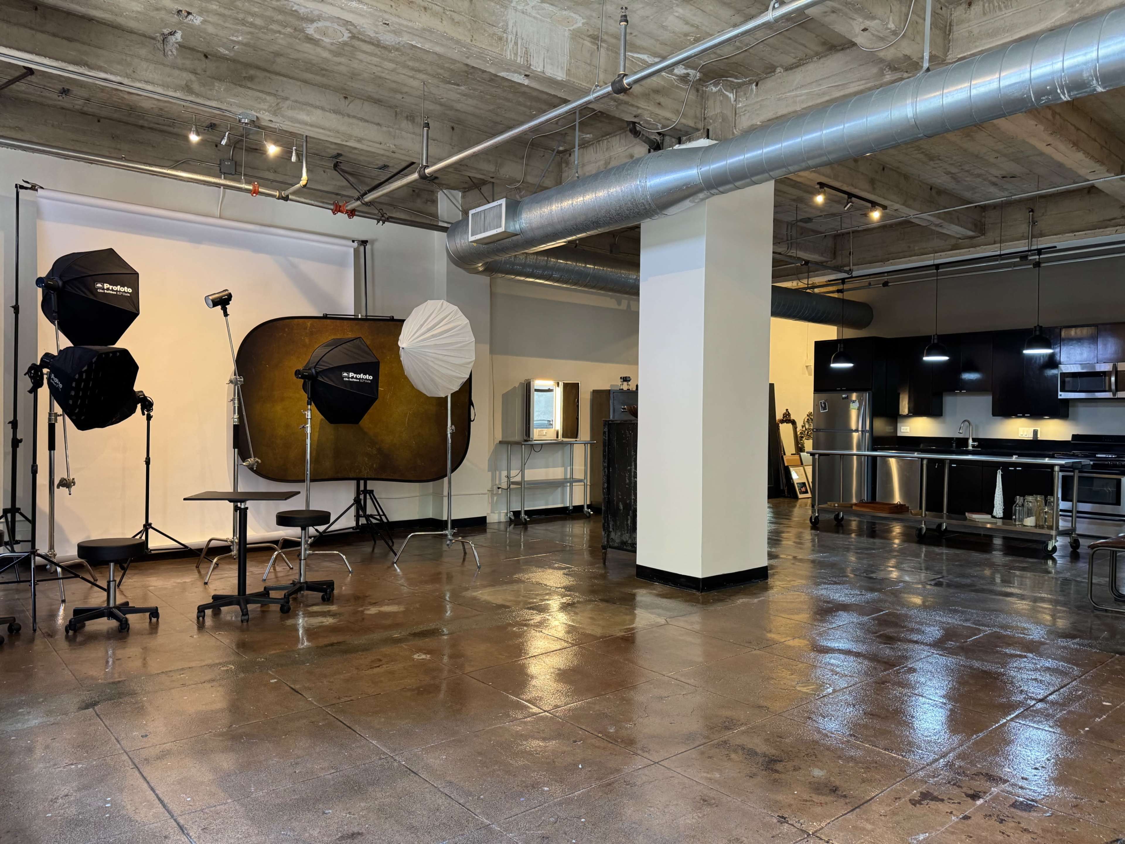 The image shows a spacious photography studio featuring various lighting equipment, a background setup, and an adjoining kitchen area.