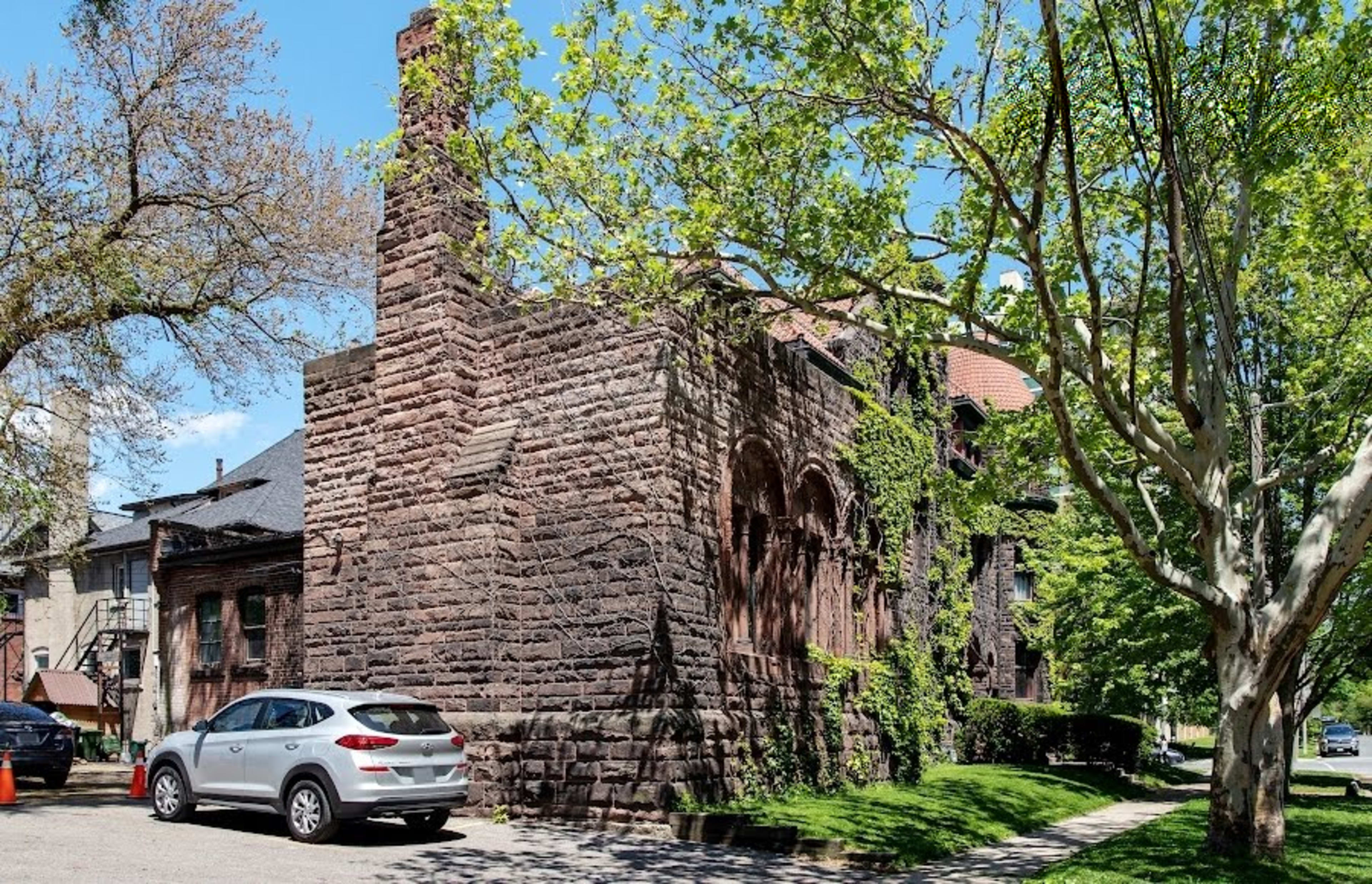 The image shows a large, stone building with a partially visible chimney, surrounded by trees and parked cars on a residential street.