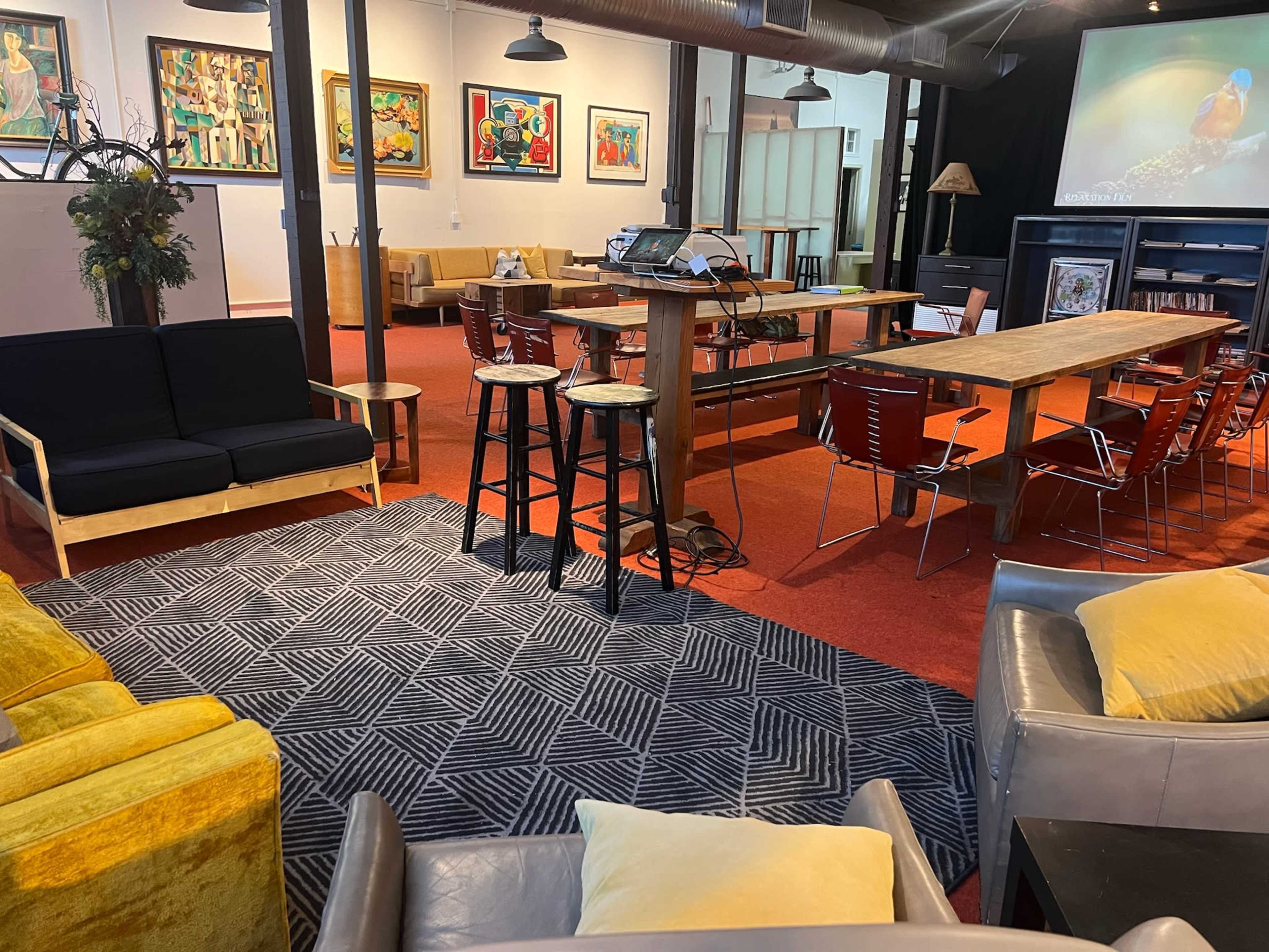The image shows a spacious interior with various seating arrangements, including sofas and chairs, alongside a wooden table and artwork displayed on the walls.