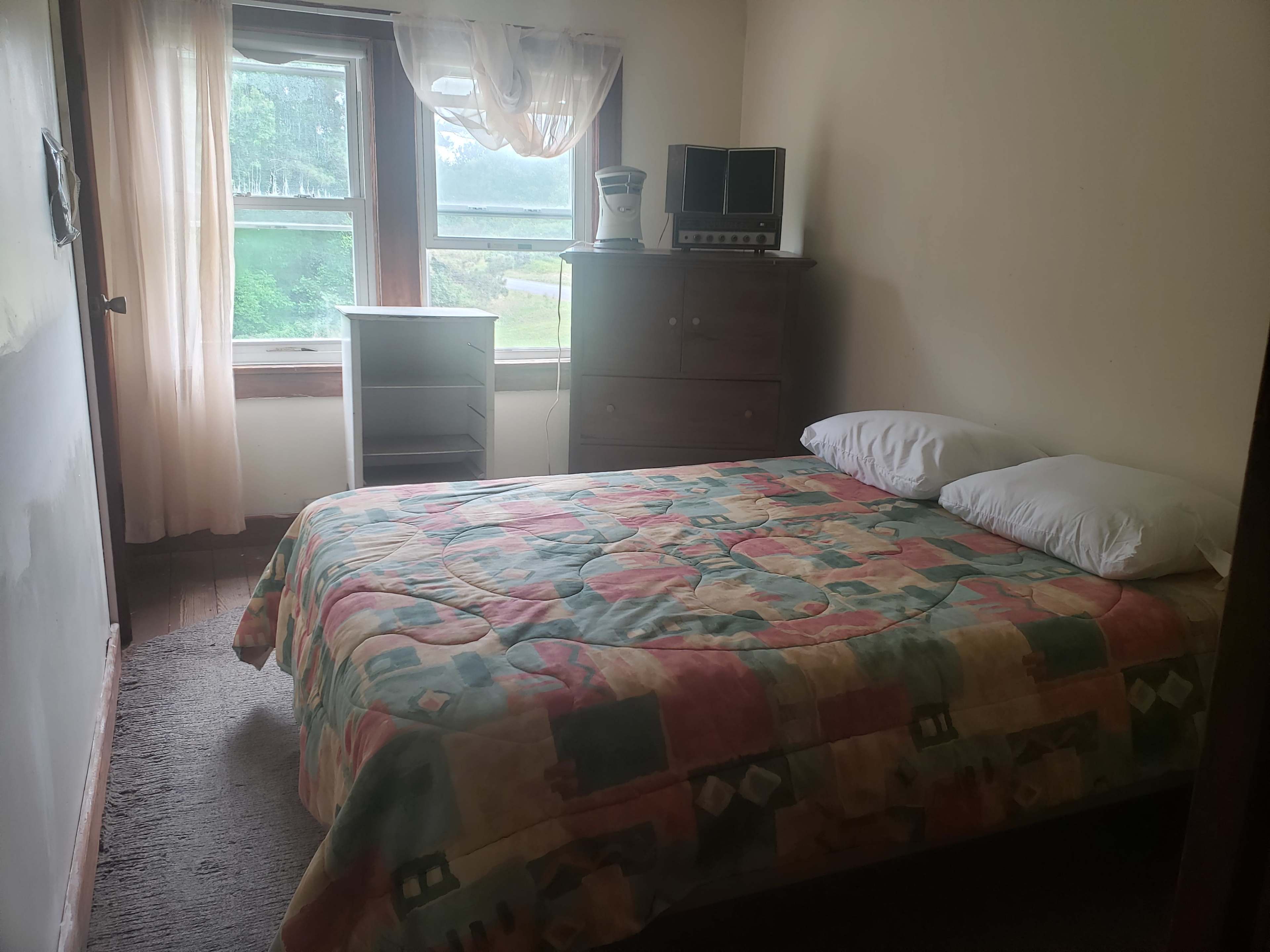 A small bedroom features a bed with a patterned quilt, a dresser, a television, and a window with sheer curtains allowing natural light.