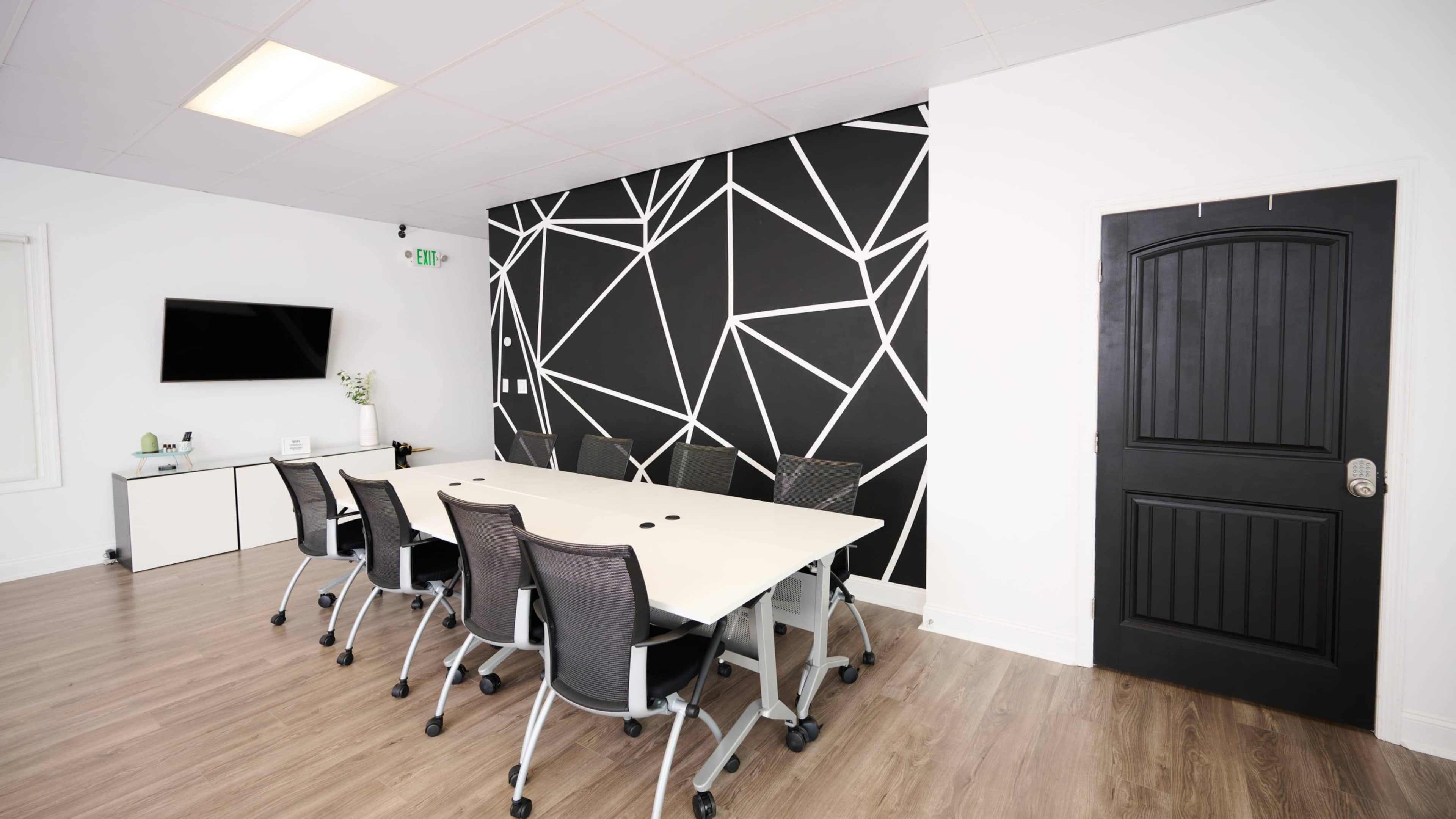 The image shows a modern conference room with a long white table surrounded by black mesh chairs, featuring a geometric black and white accent wall and a TV on one side.