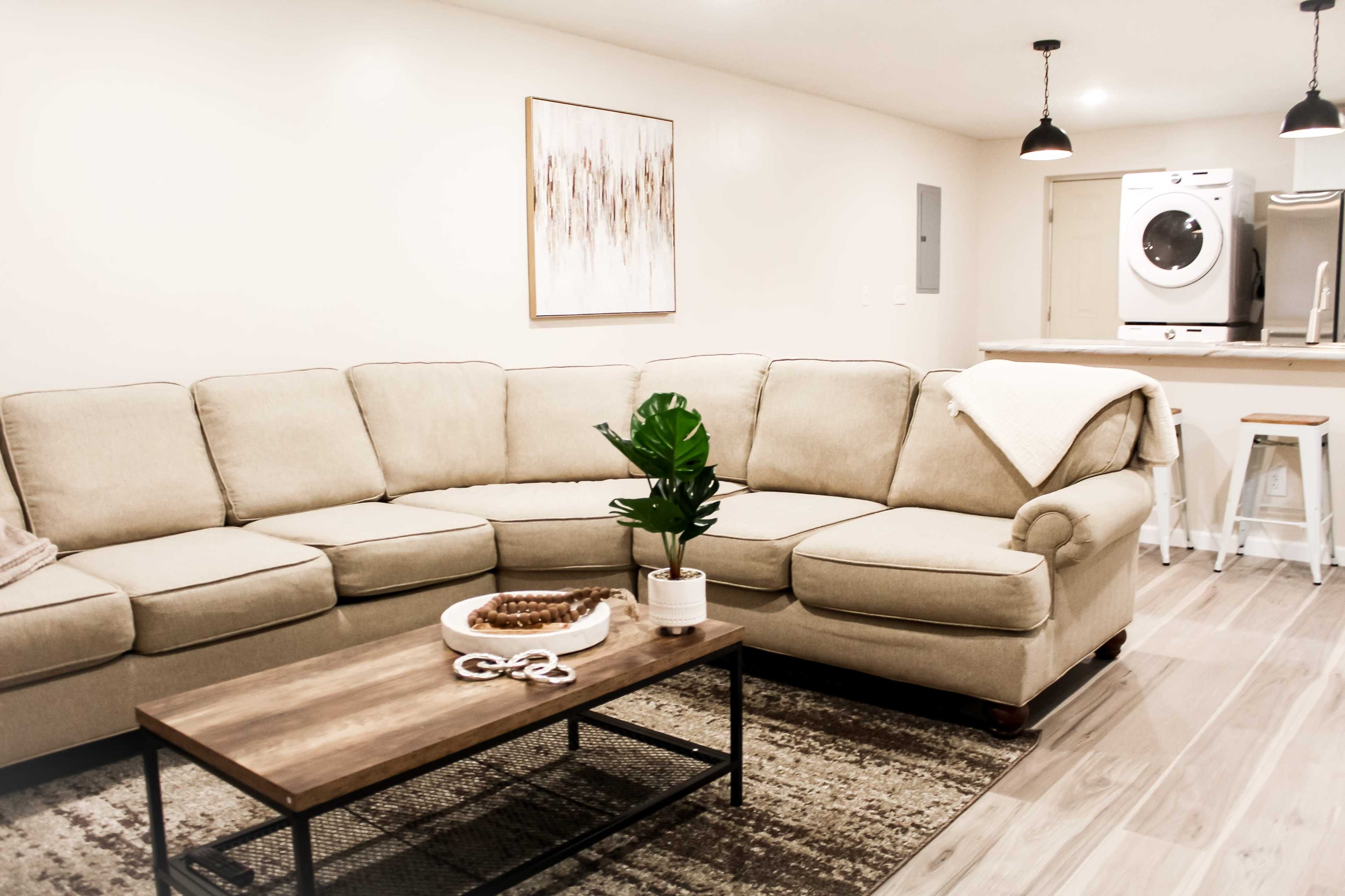 A beige sectional sofa is positioned in a living room with a wooden coffee table, a small potted plant, and a laundry area visible in the background.