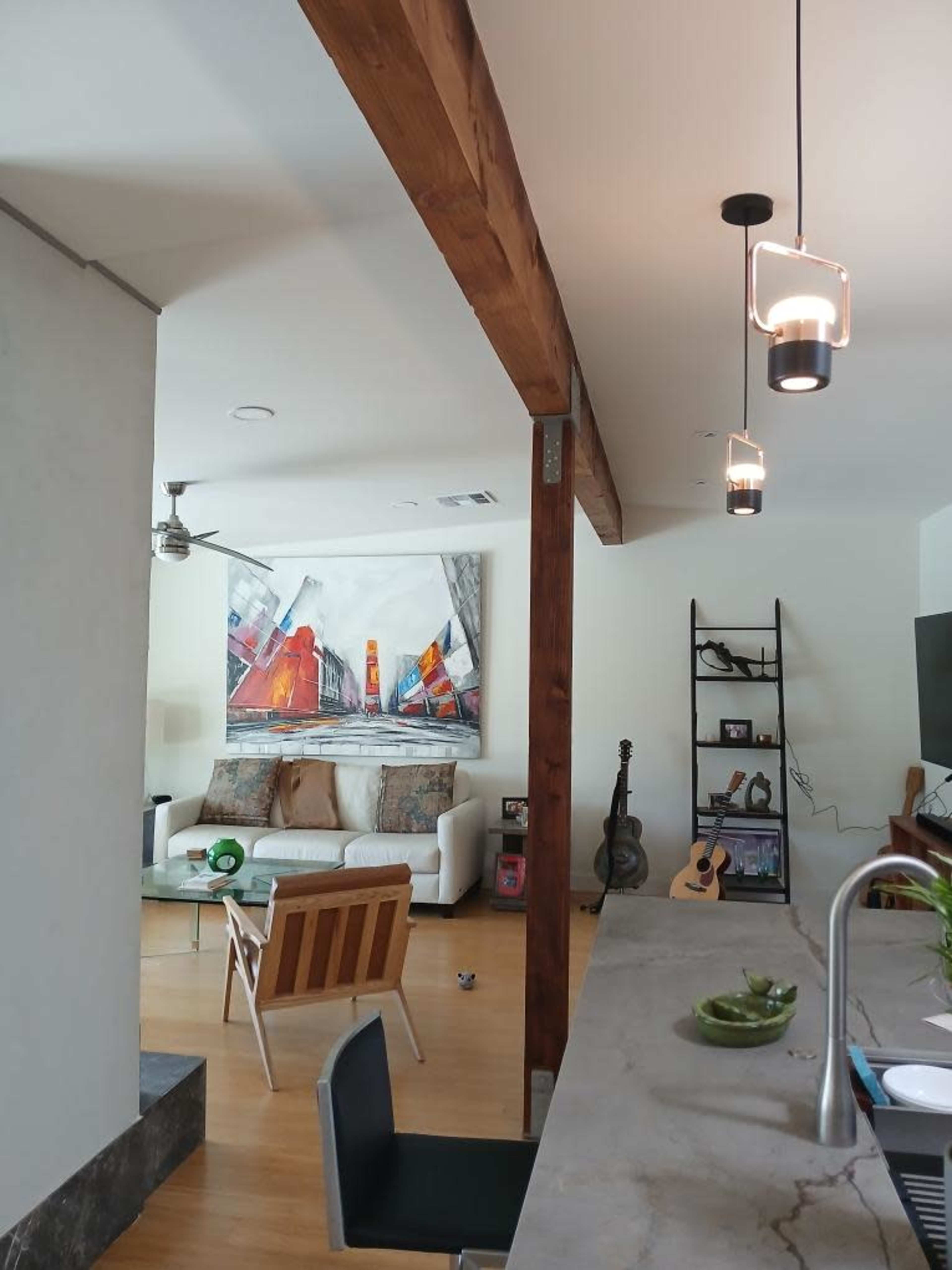 The image shows a modern living space featuring a couch, a large abstract painting, and a kitchen island with a sink and plants.