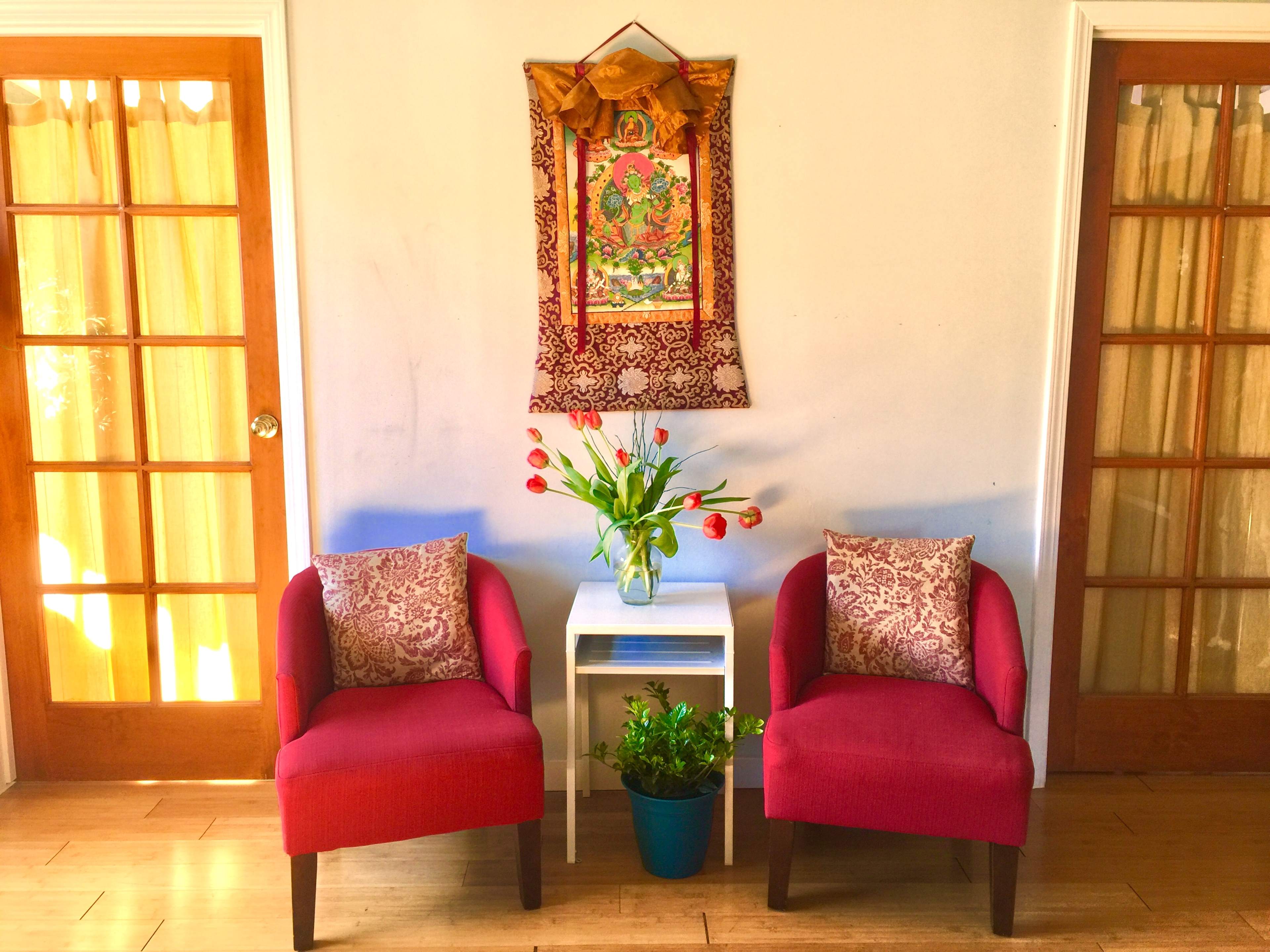 Two red chairs with decorative pillows are positioned on either side of a small white table, beneath a colorful wall hanging, with a vase of tulips and a potted plant in view.