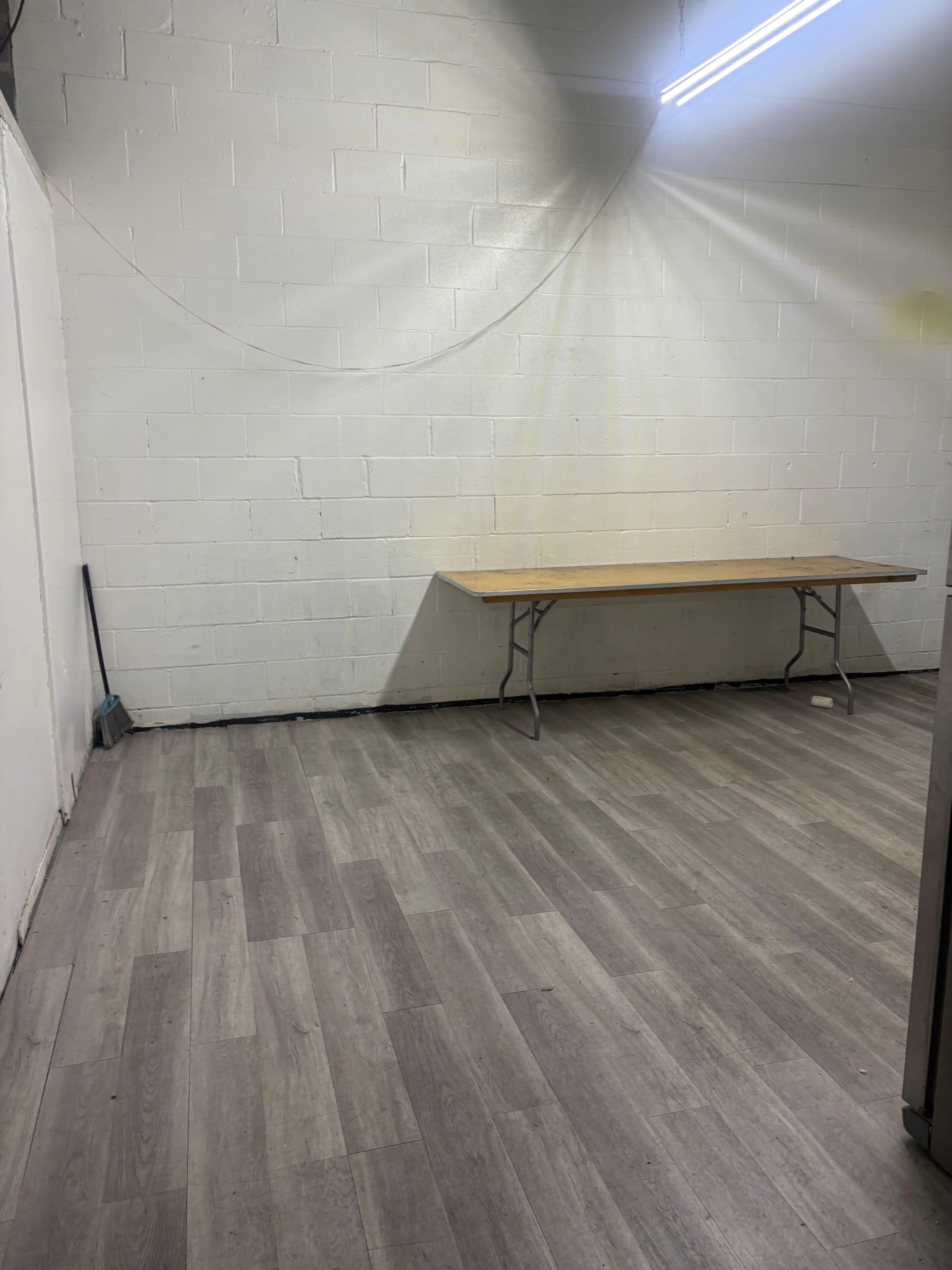 The image shows a bare room with a white concrete wall, a wooden table against one side, and a floor with light gray laminate.