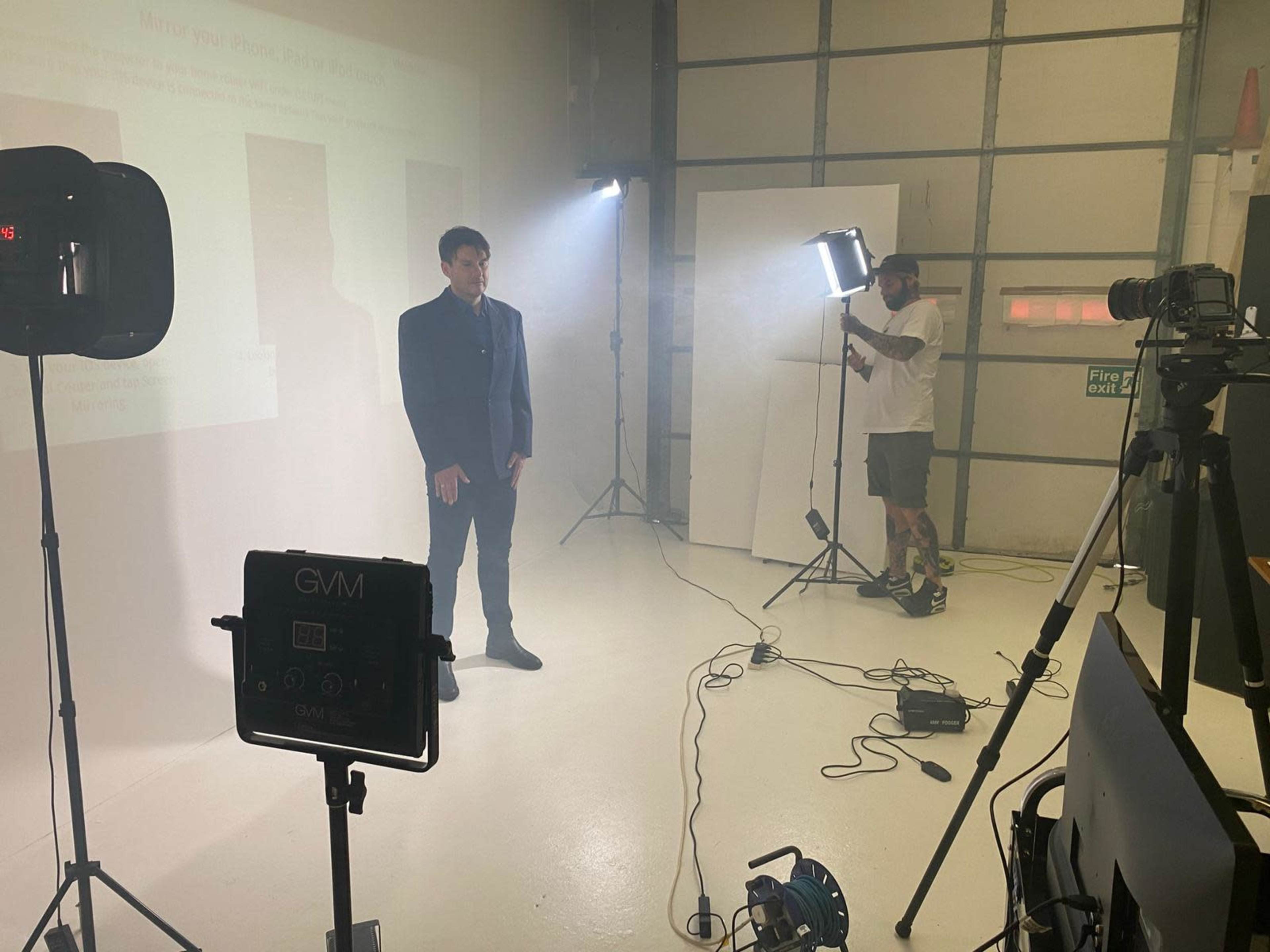 A man in a suit stands in a studio surrounded by lighting equipment and a camera operator.