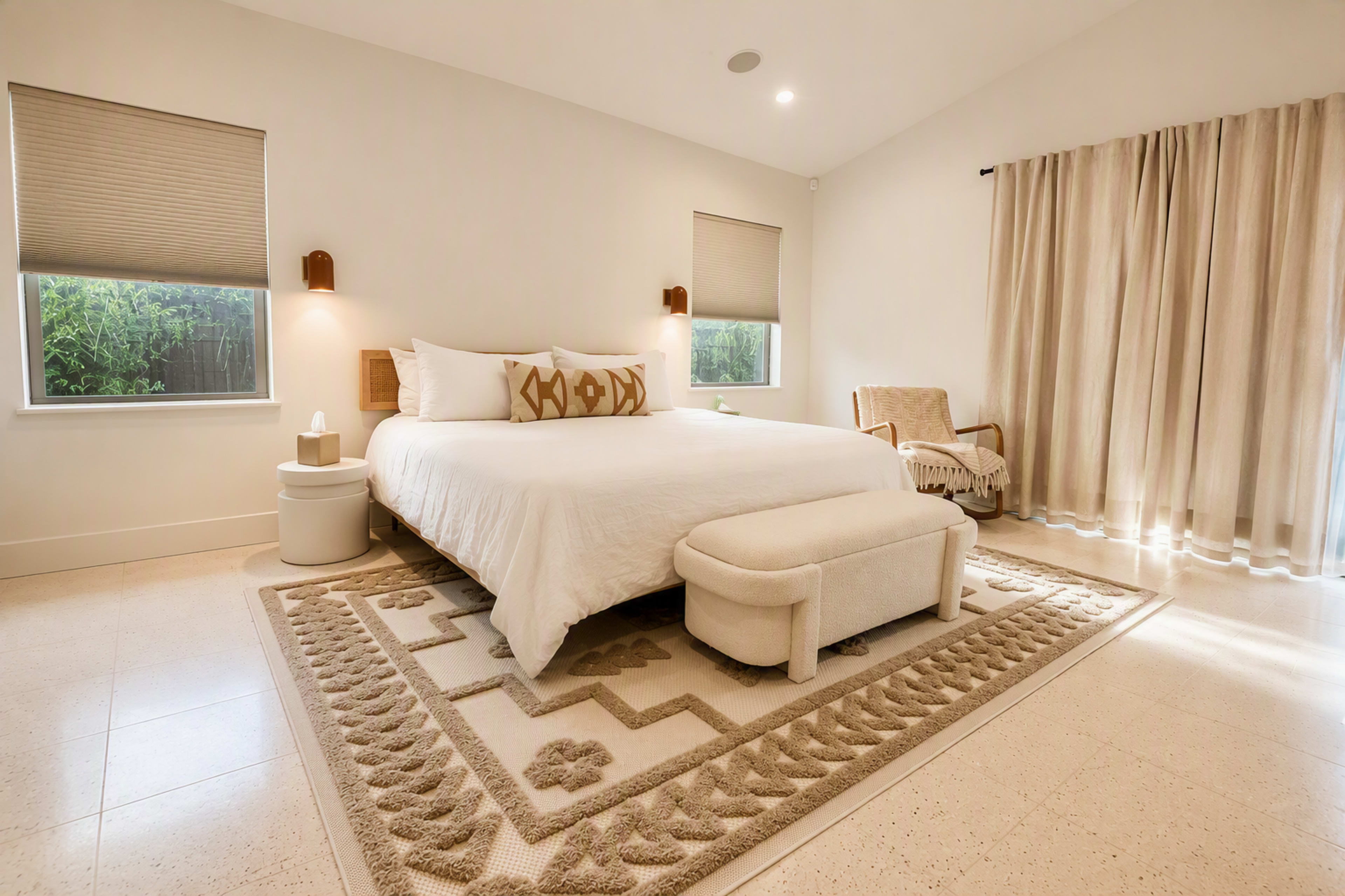 The image shows a spacious, well-lit bedroom featuring a large bed with decorative pillows, a plush bench at the foot, and natural light streaming through the windows.
