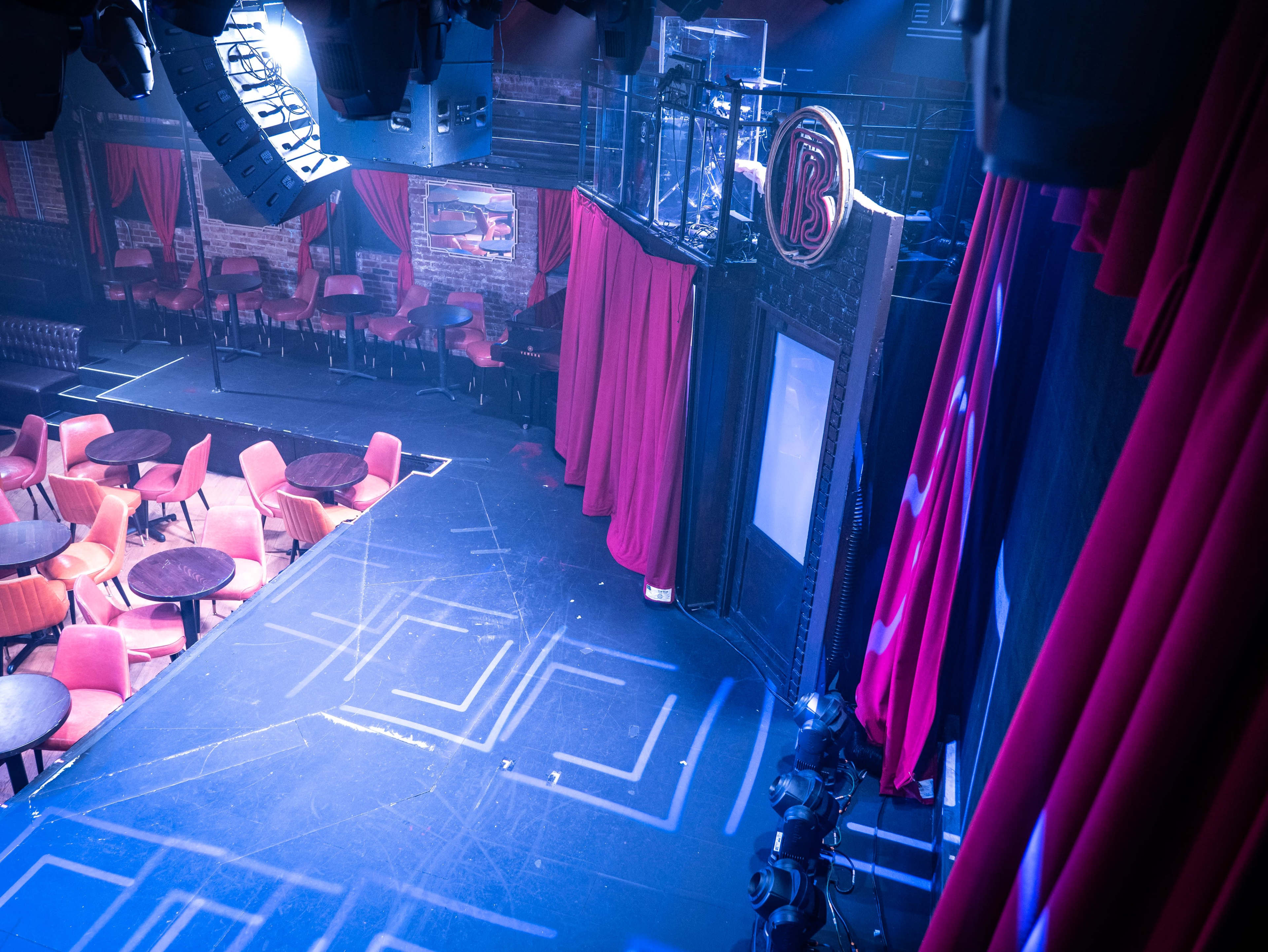 A dimly lit stage with red curtains, scattered tables and chairs, and a spotlight setup indicating a performance area in a theater.